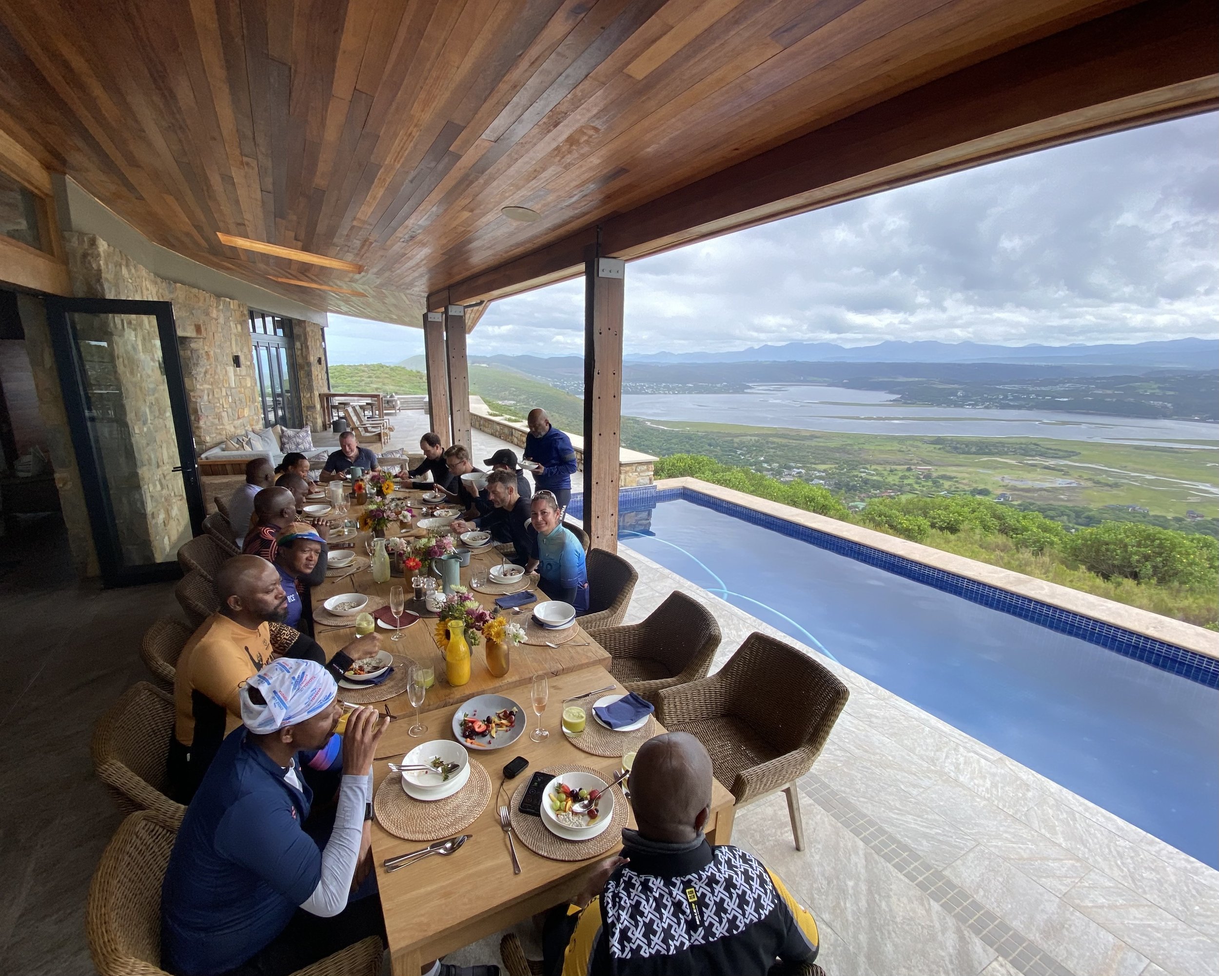 People dining together at an outdoor restaurant with a scenic view of a river and mountains in the background. The setting features a wooden ceiling and stone walls. The table is set with plates, glasses, and flowers.