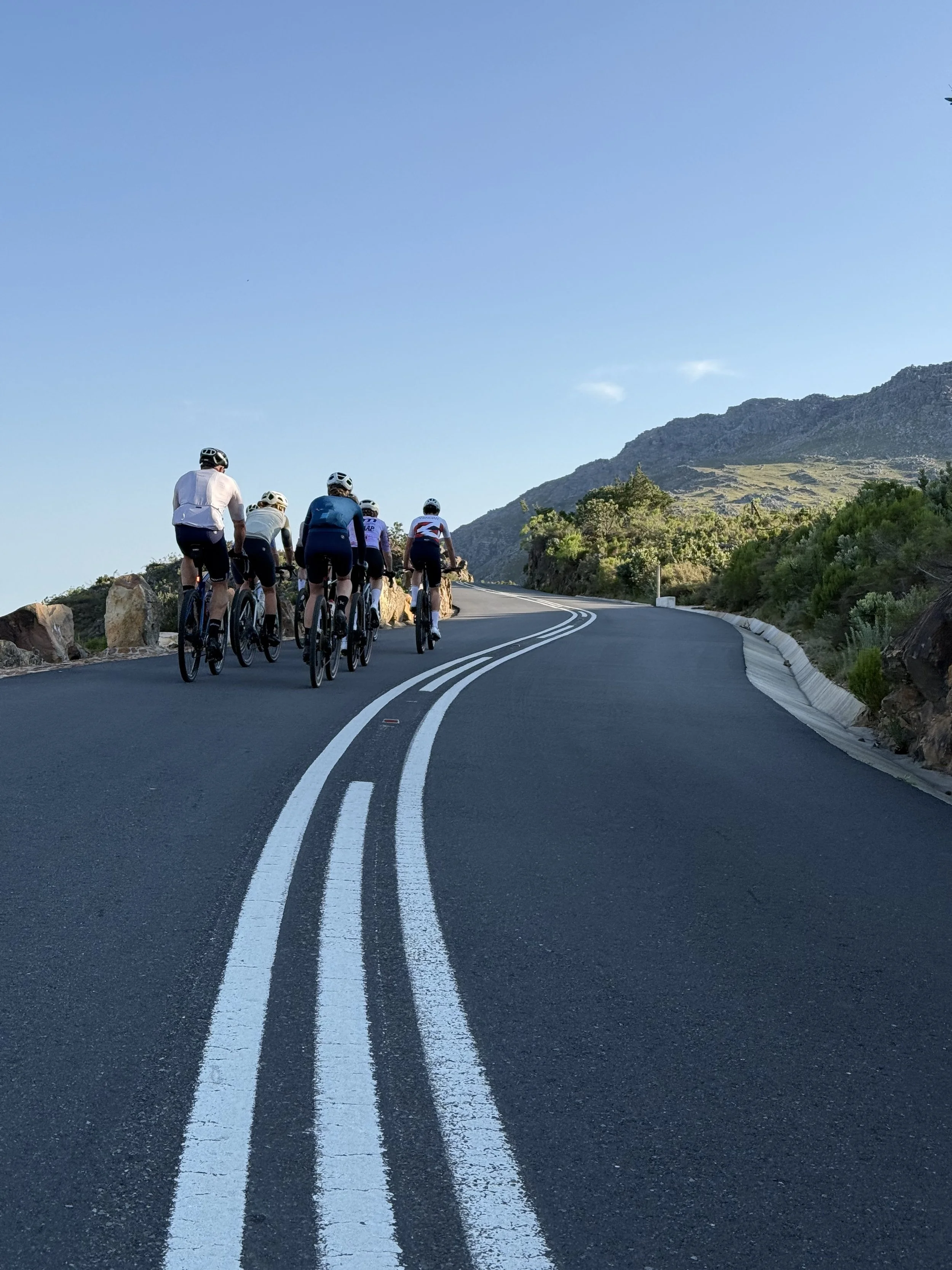 A group of cyclists riding up a winding mountain road on a clear day with a blue sky and surrounding greenery.