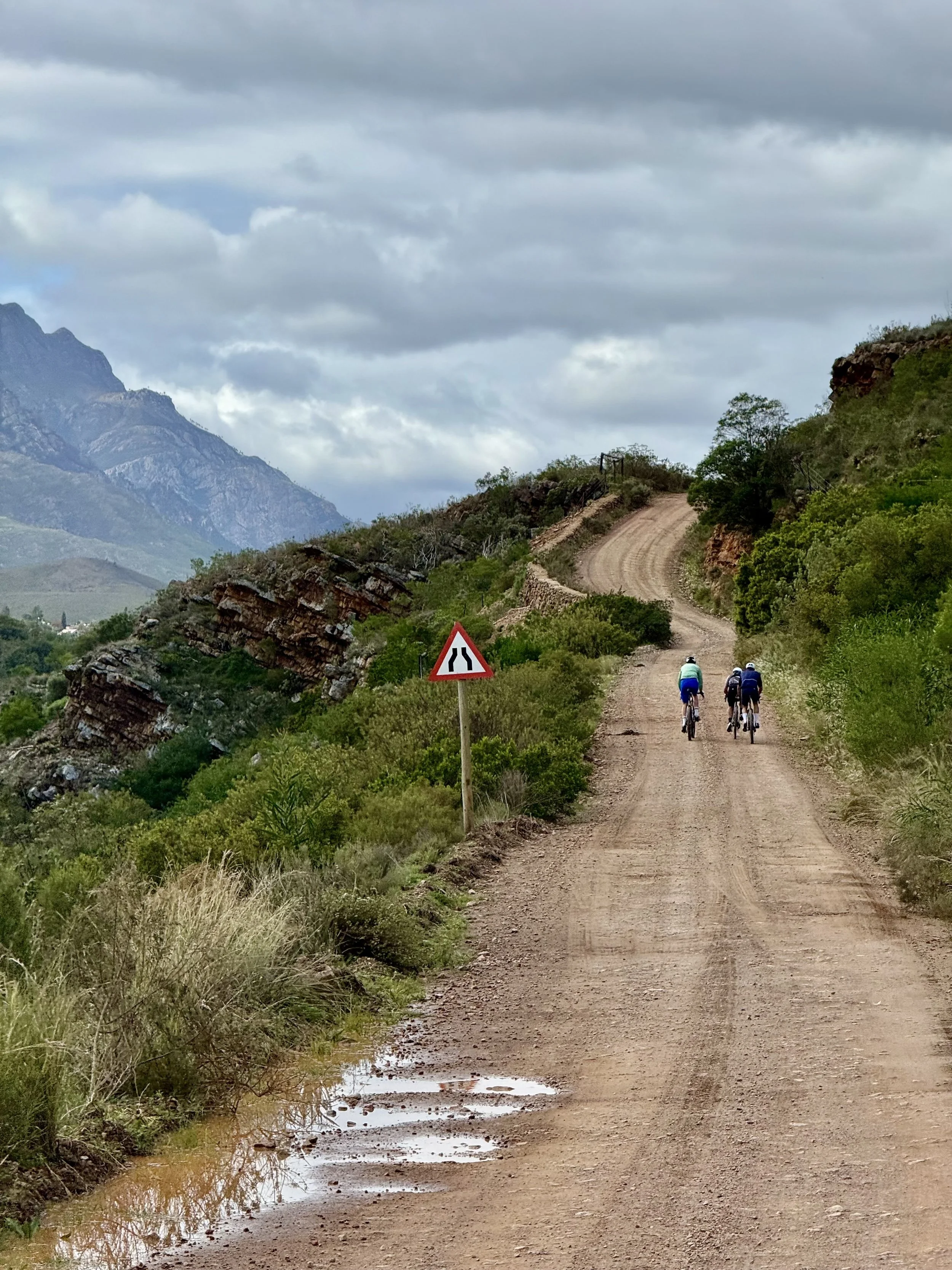 A dirt road winding through a hilly landscape with green bushes and trees, mountains in the background, and a cloudy sky overhead. Three cyclists are riding along the road, and there is a traffic sign indicating a narrowing road.
