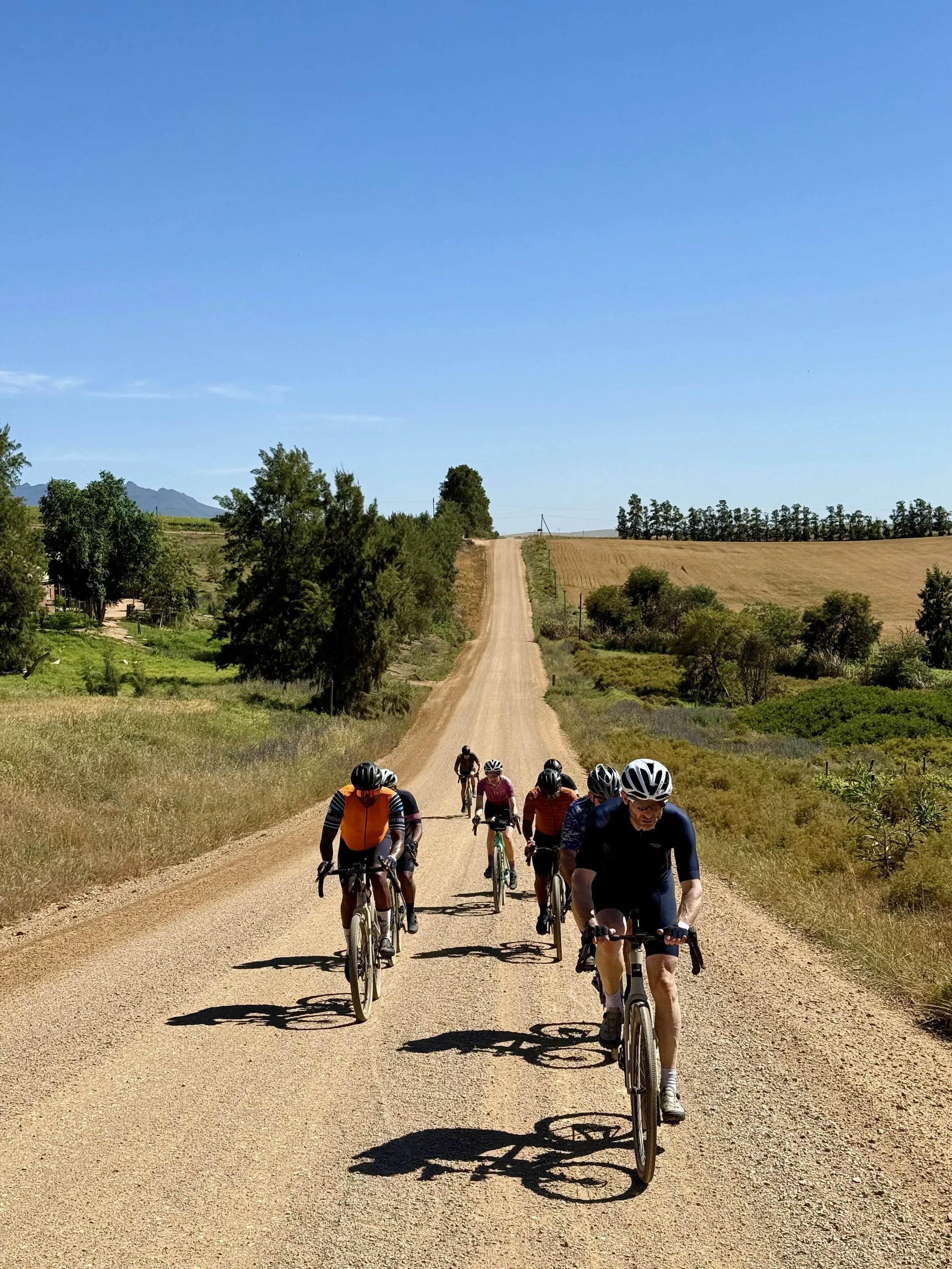 A group of cyclists riding on a dirt road through a rural landscape with green trees and fields under a bright blue sky.