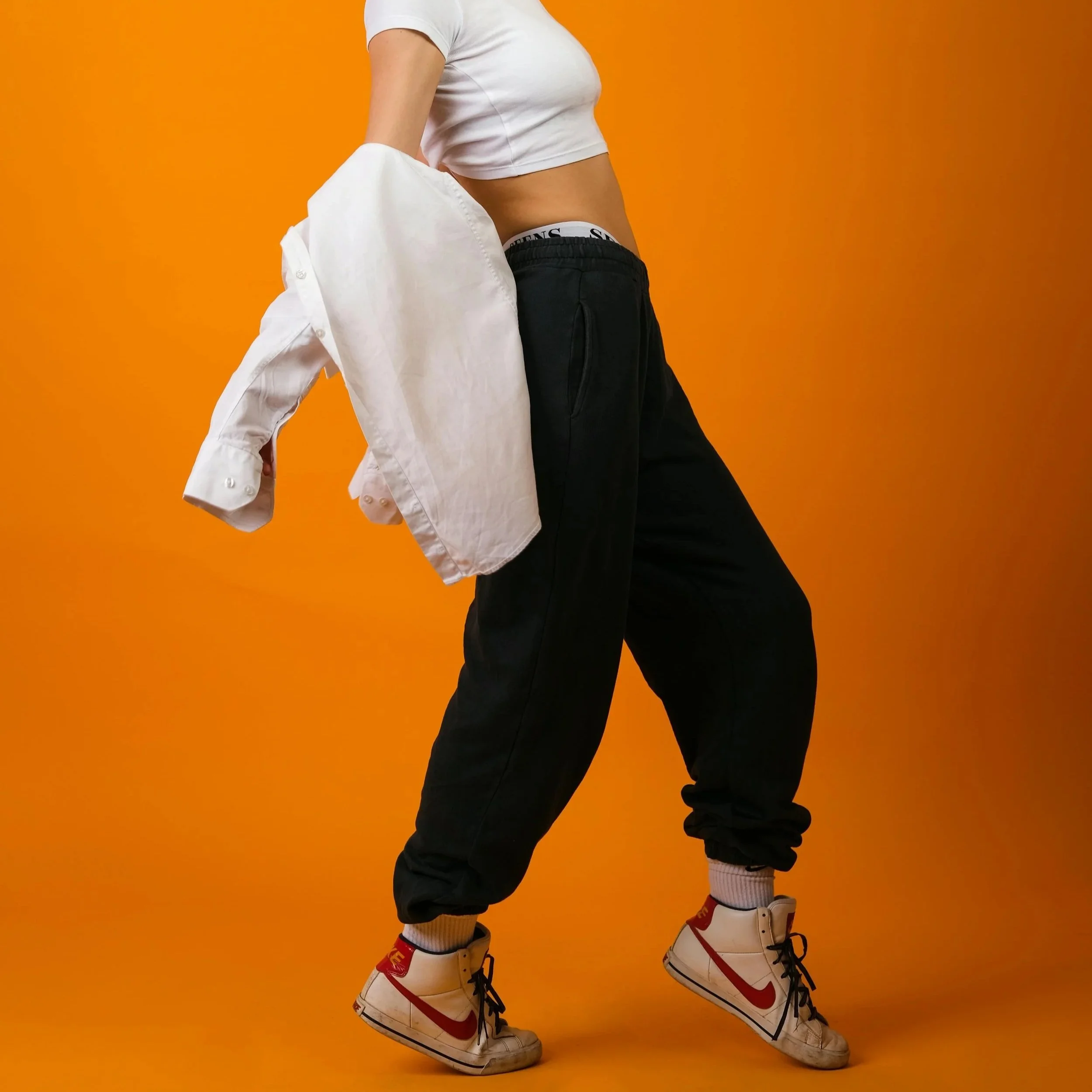 A person wearing white and red Nike sneakers, black joggers, a white crop top, white socks, and a white shirt draped over their shoulder, standing against an orange background.