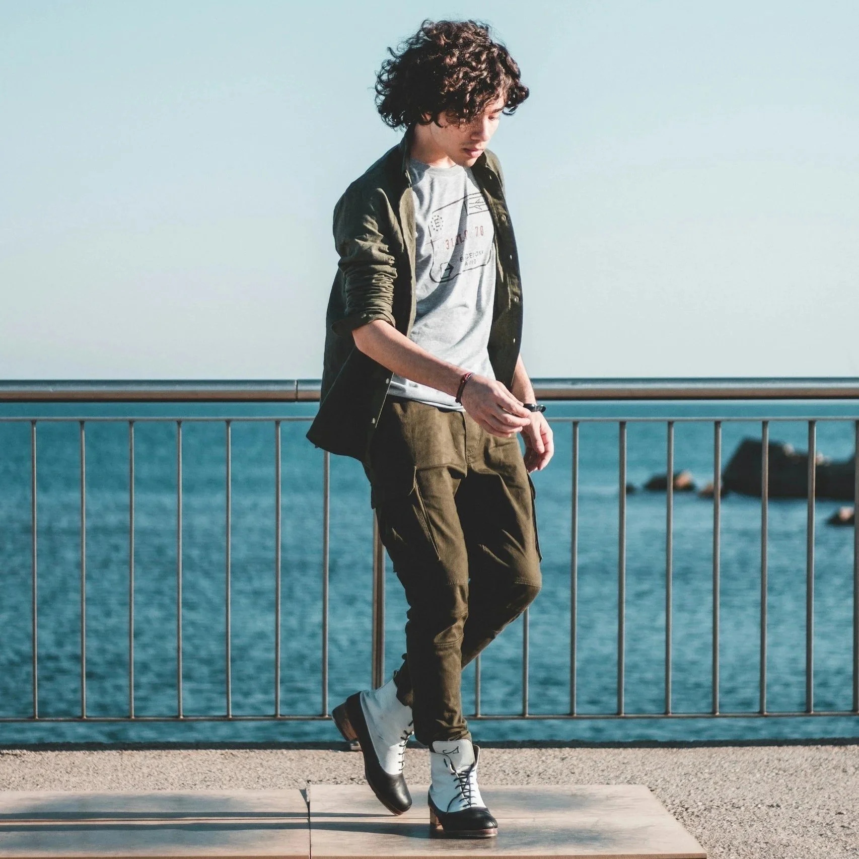 Young man with curly hair standing on a sidewalk near a metal railing by the ocean, wearing a green jacket, gray t-shirt, khaki pants, and white and black boots, looking down with a contemplative expression.