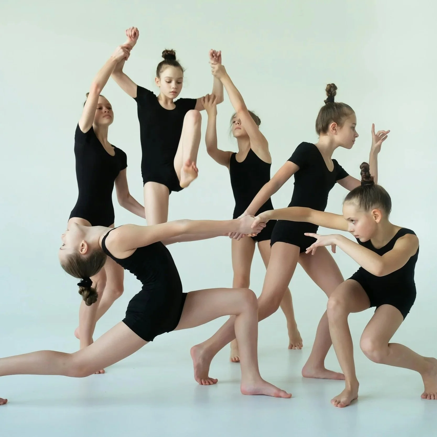 A group of young women dance in black leotards in a studio, forming a complex human sculpture with various poses and handshakes.