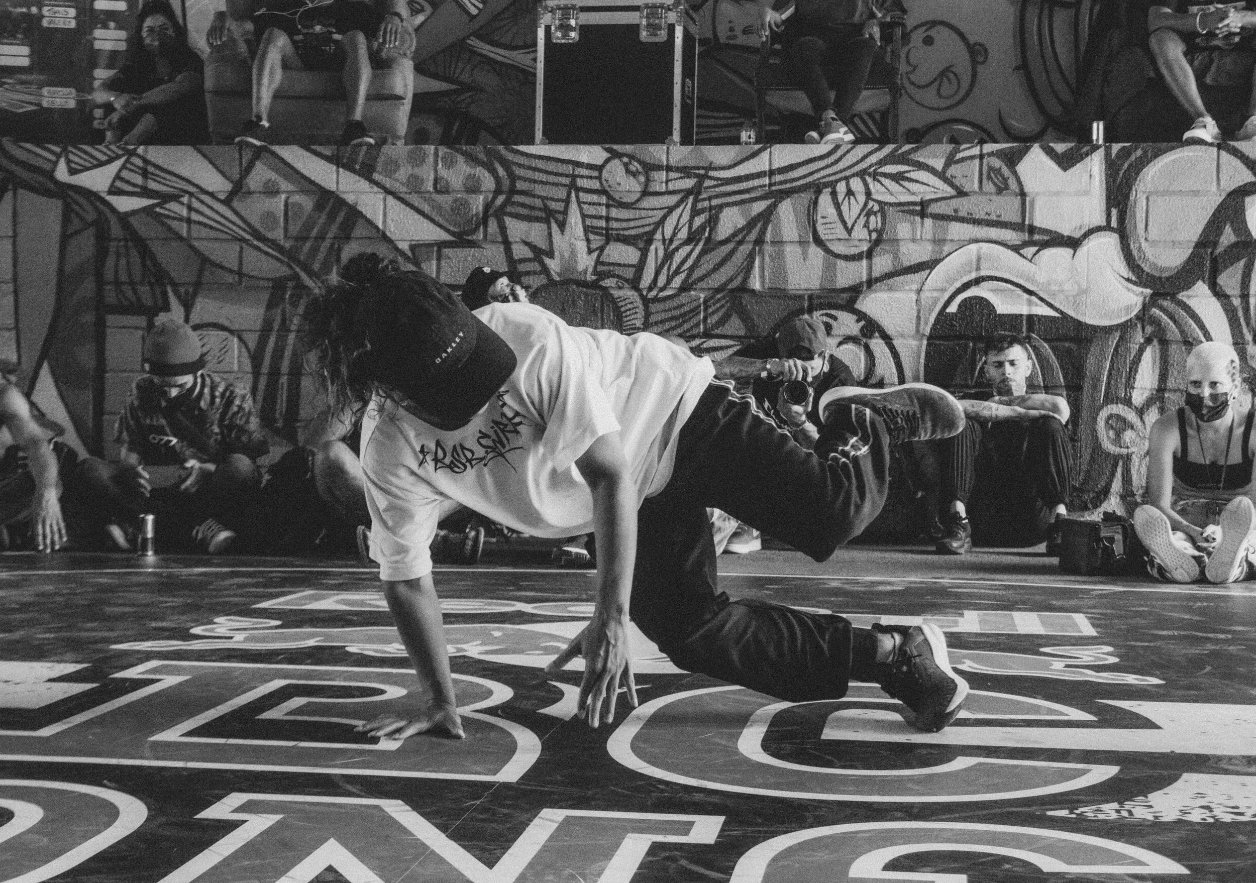 A group of people and a performer are in front of a graffiti-covered wall, with some people sitting and some standing, some watching and some taking photos. A man is in the foreground performing a dance move on the floor.