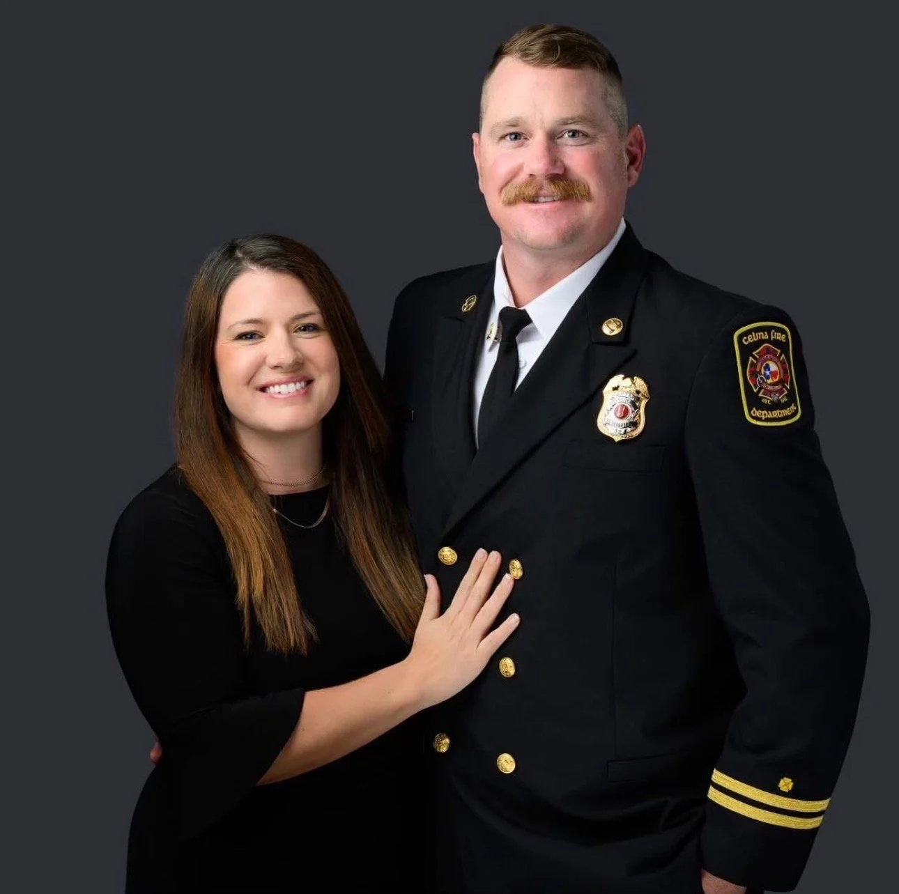 A woman with long brown hair and a man in a firefighter uniform pose together against a dark gray background, smiling.