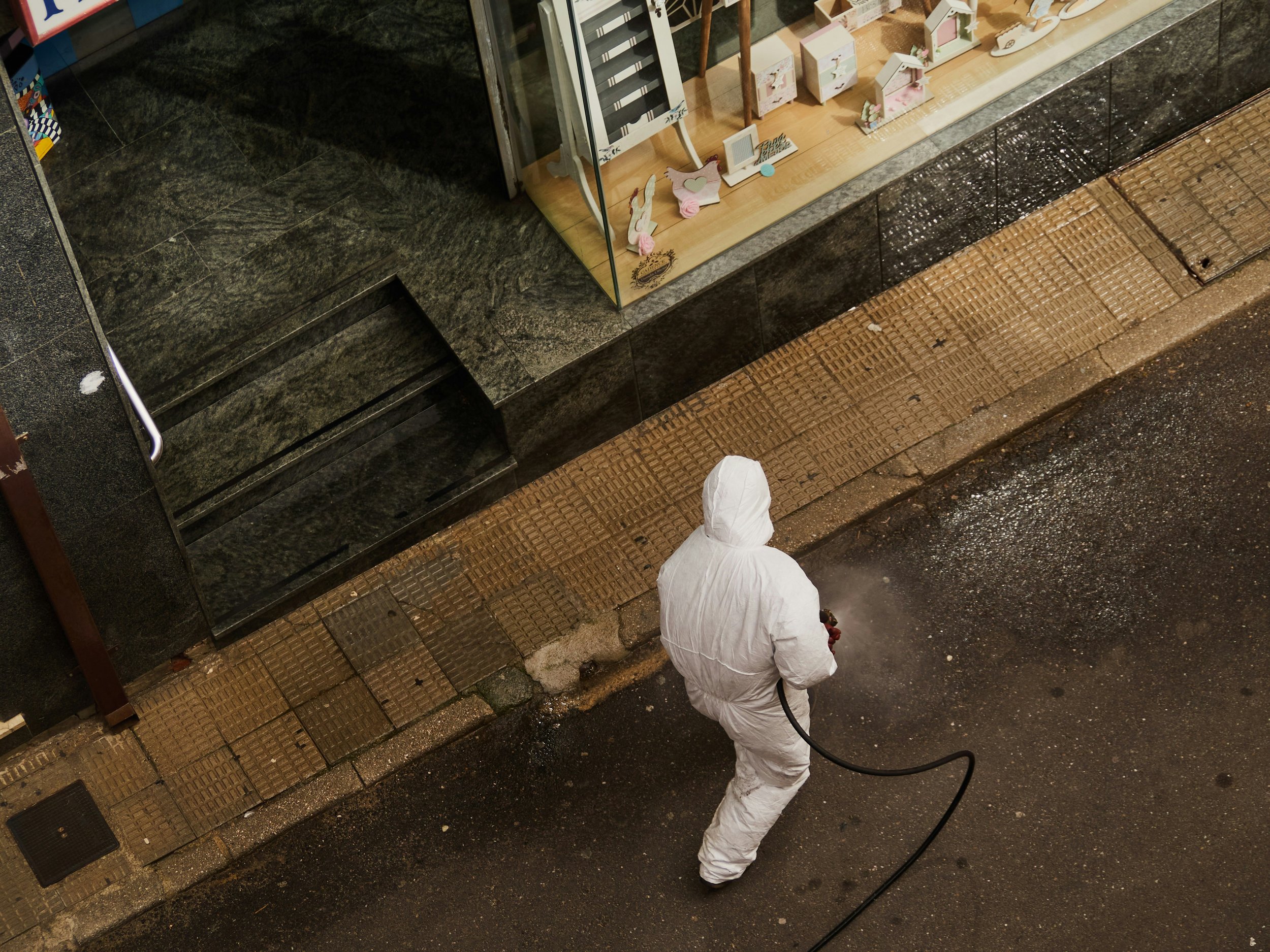 A person in a white protective suit with a hood, standing on a wet sidewalk at night, holding a black hose, in front of a store display window with decorative items.