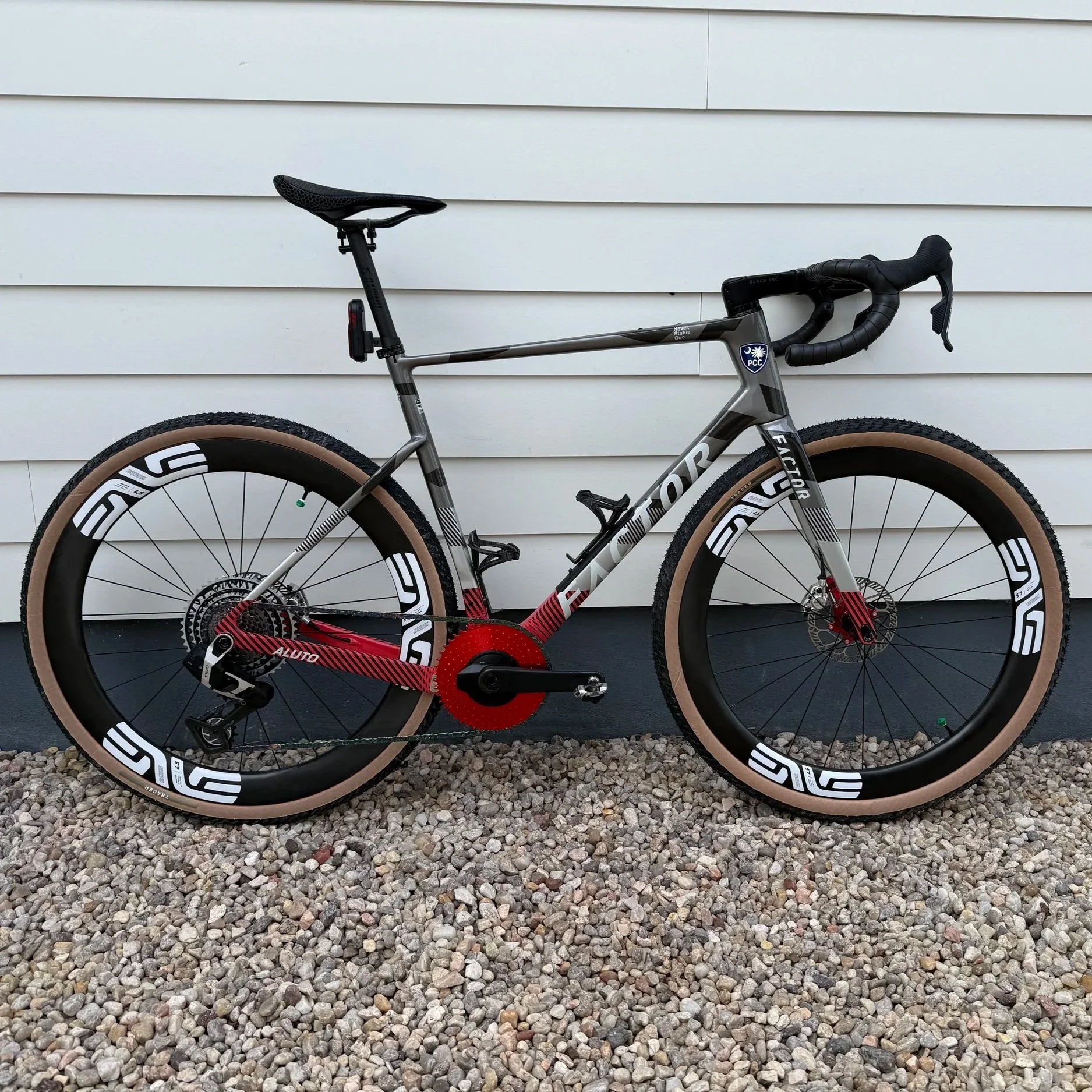 A modern road bike with a grey and black frame, black handlebars, black saddle, and tan tires, leaning against a white horizontal siding wall on a gravel ground.