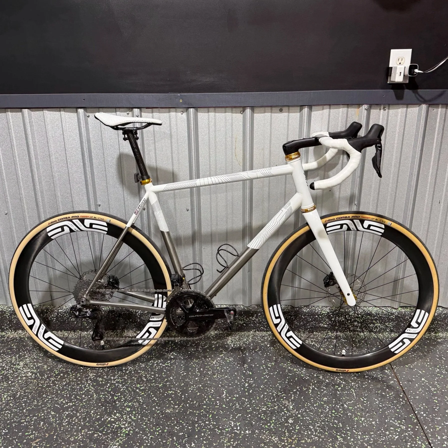 A white road bike with black and gold accents resting against a metal wall in a gym or storage area with a black wall and electrical outlets.