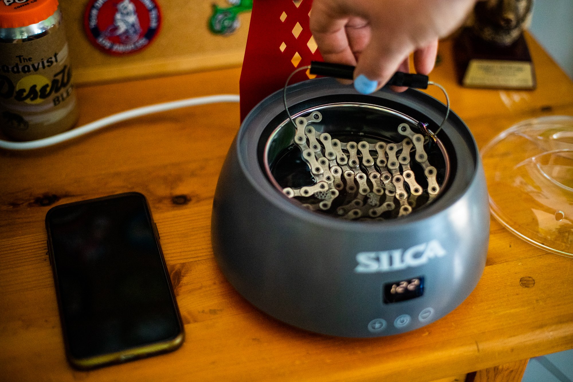 A person removing a nail from a electric nail polisher machine using a black tool. The machine is placed on a wooden table with a smartphone and other objects nearby.