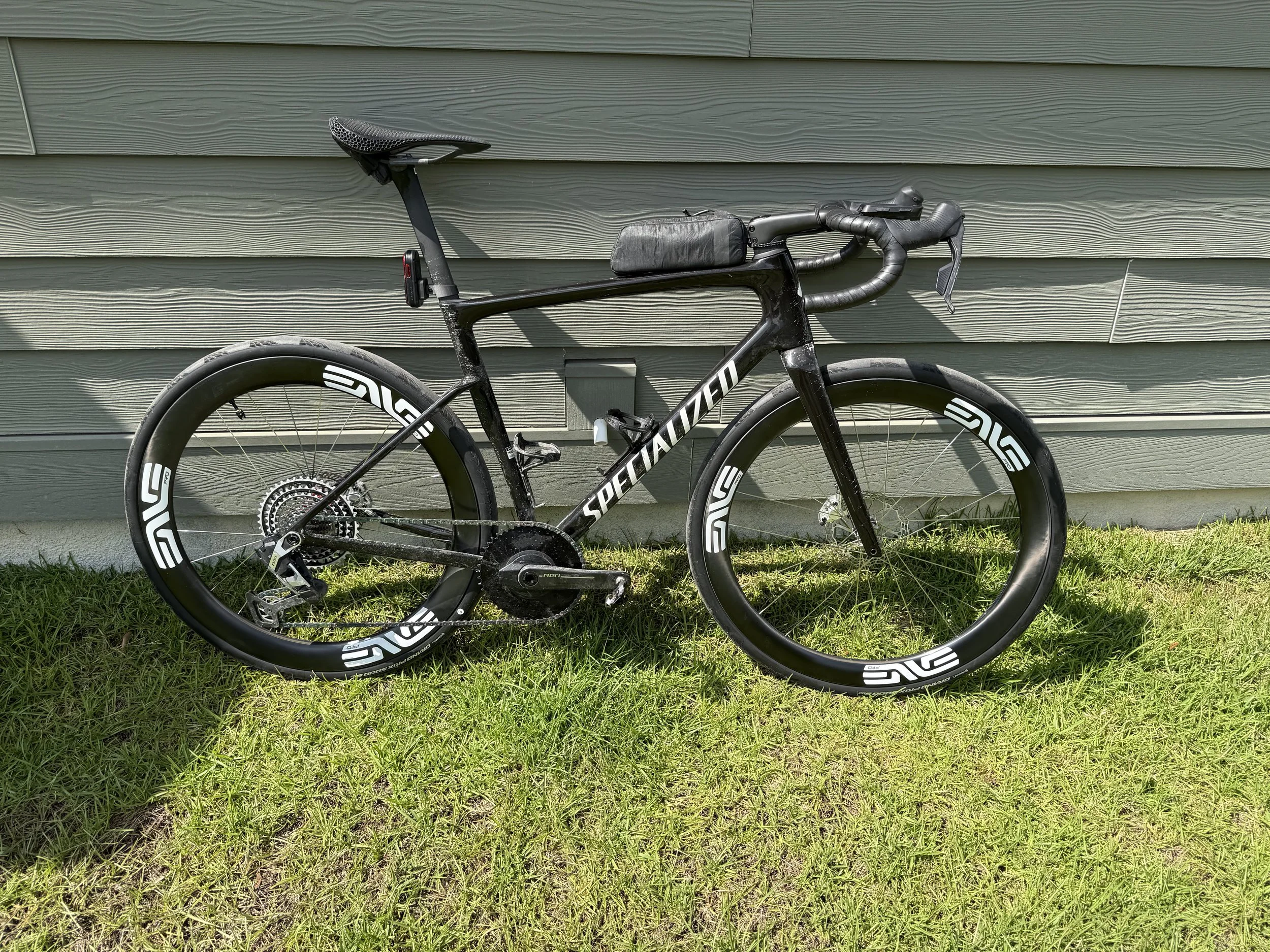 A black Specialized brand road bicycle leaning against a gray house siding wall with grass below.