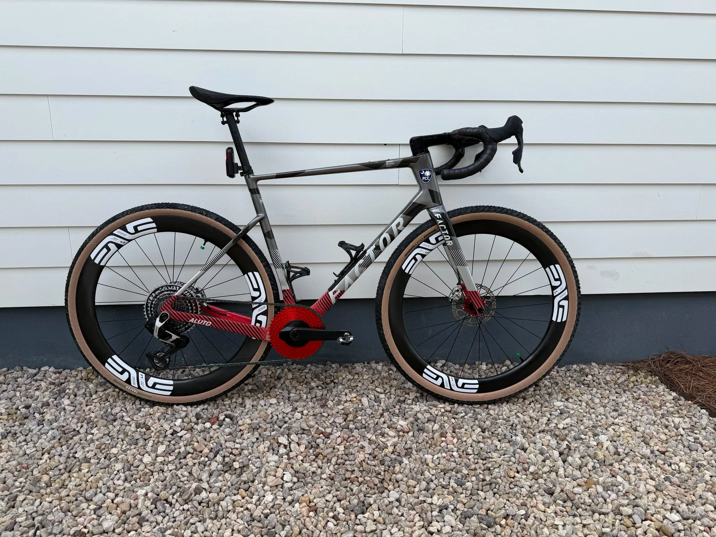 A gray and black racing bicycle with beige tires is positioned against a white vinyl-sided wall, resting on a gravel ground.