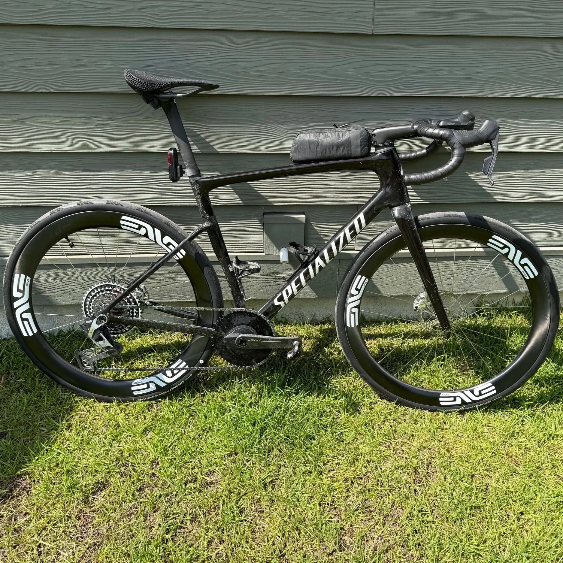 A black Specialized S-Works road bike with disc brakes, aerodynamic wheels, and handlebar gear shifters resting on green grass against a gray house siding background.