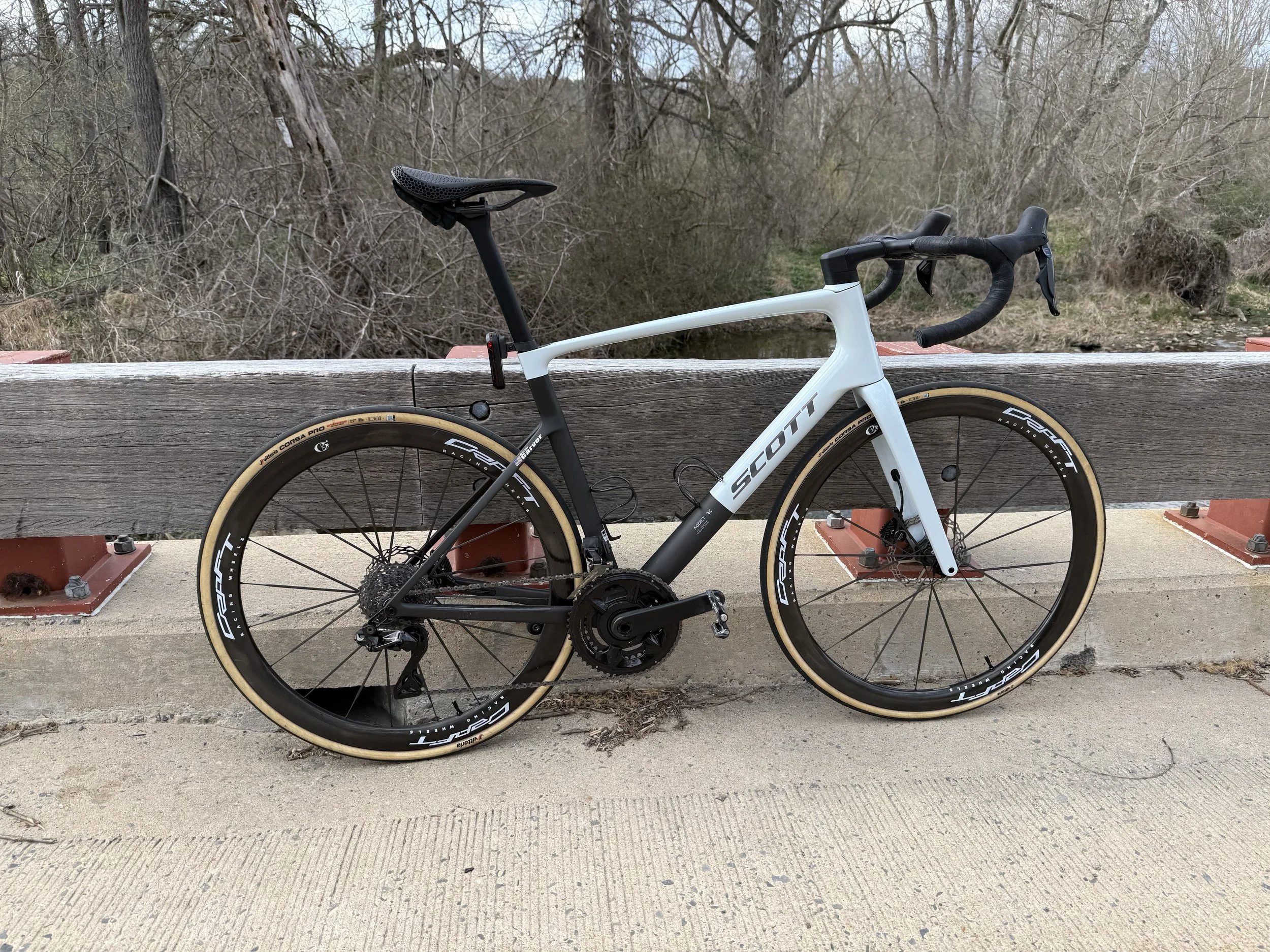 A white road bicycle with black and tan tires parked against a wooden railing on a bridge, with leafless trees and bushes in the background.