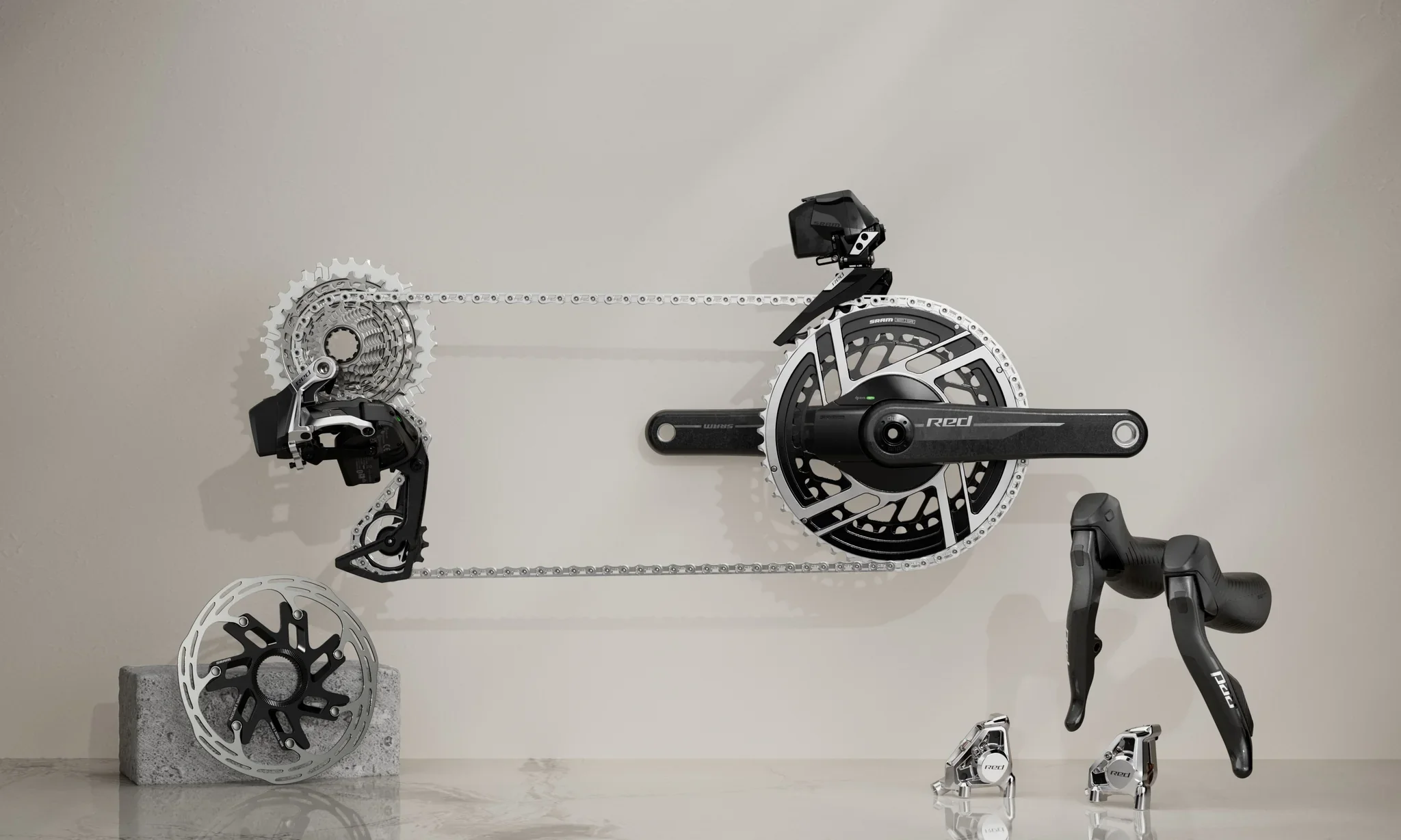 A bicycle crankset and components, including chainrings, derailleurs, and a cassette, arranged on a wall with some parts placed on a concrete block and small stands, with a game controller visible on the right.