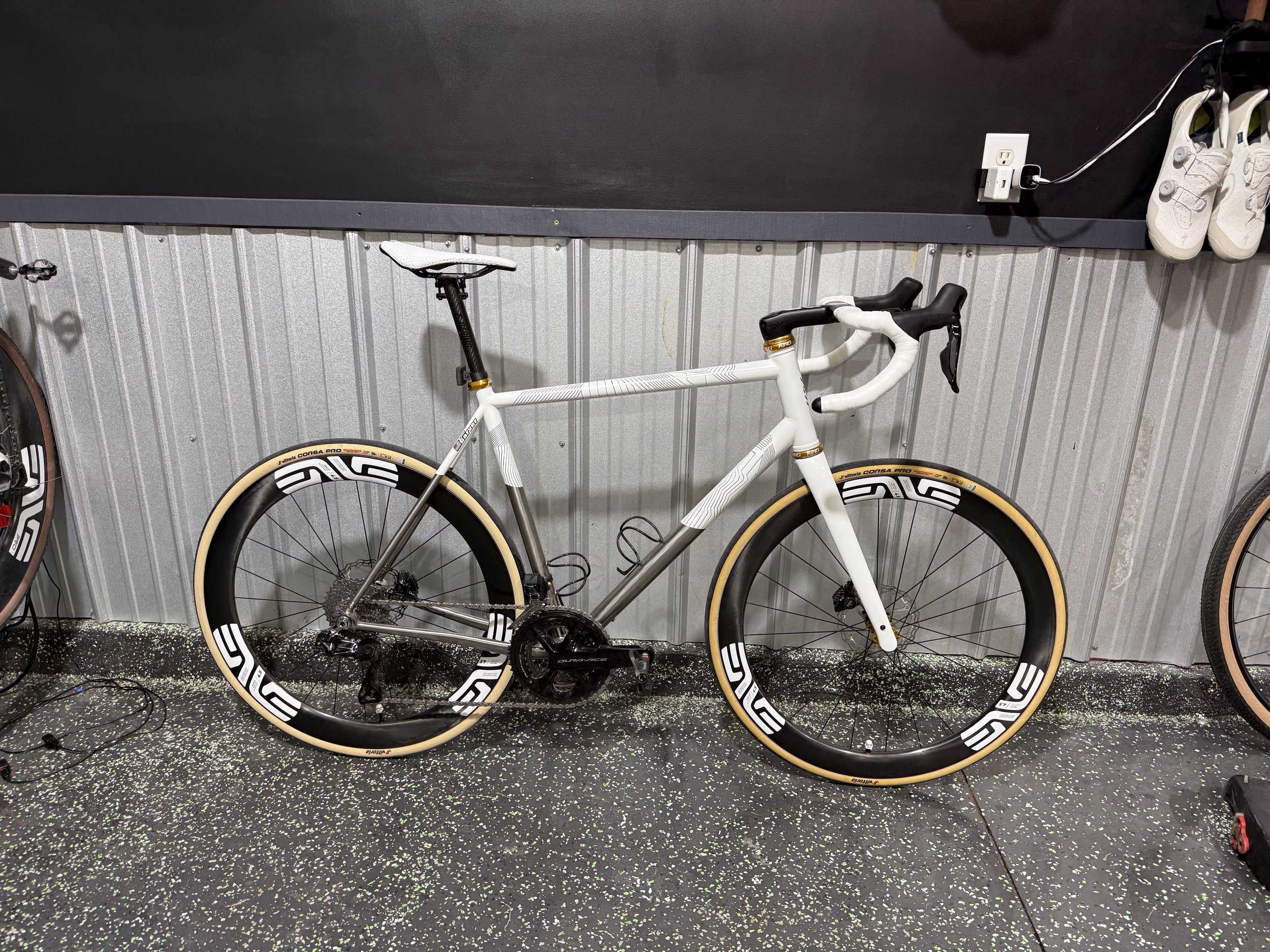 A white bicycle with black and gold accents, mounted on a black and multicolored speckled garage floor, with white sneakers and a charging cord/dock on the wall behind it.