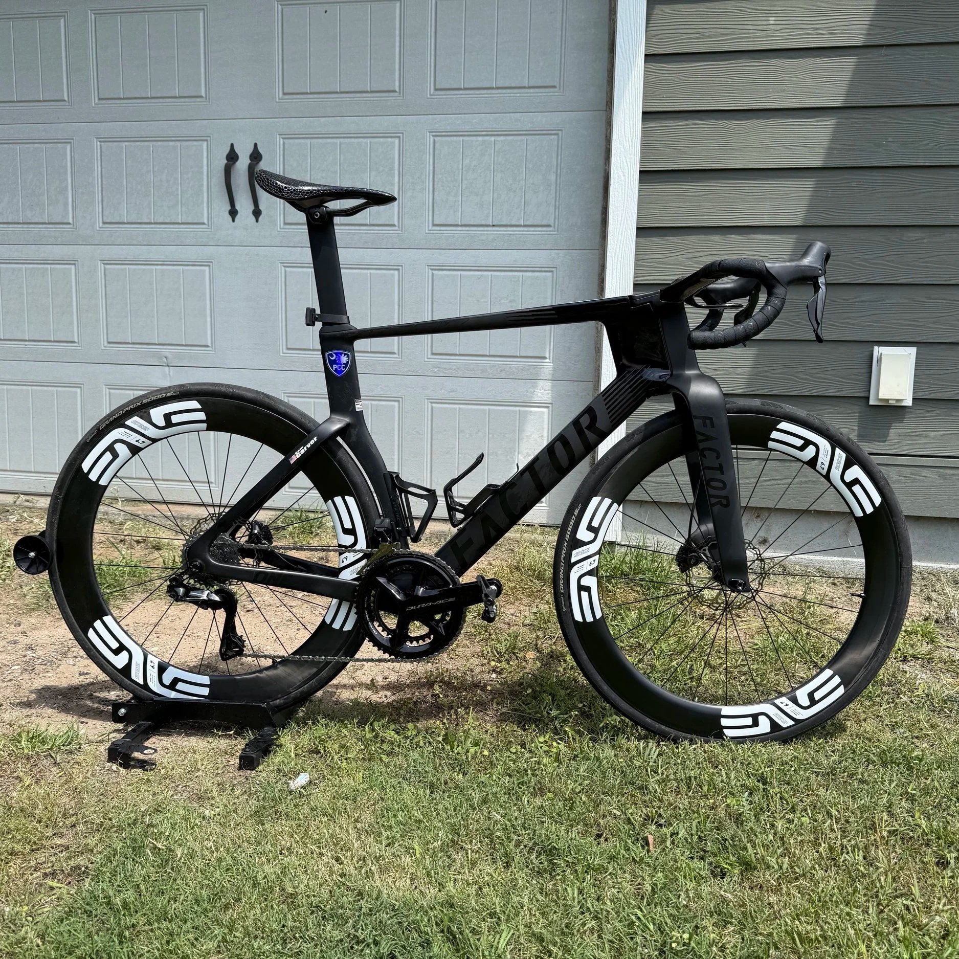 A black racing bicycle with white accents is displayed on a bike trainer outdoors, in front of a gray garage door and gray house siding, with patches of grass on the ground.