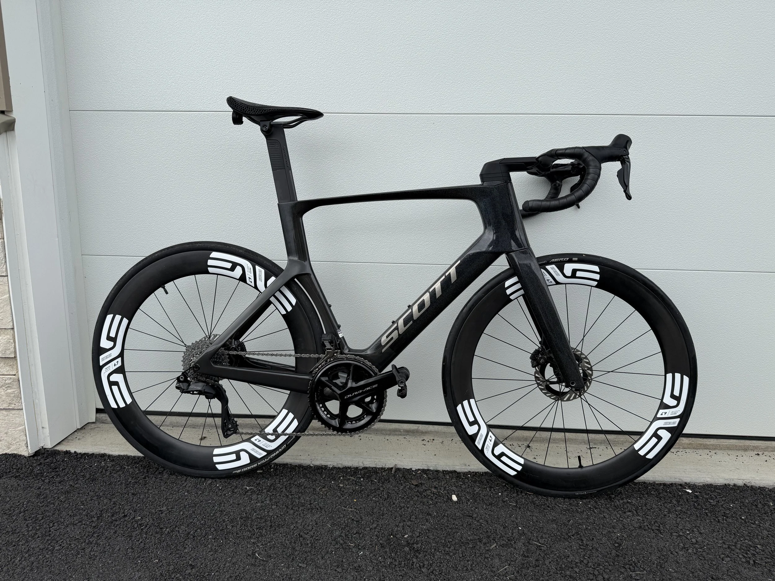 A black road bicycle with white and black racing wheels is parked against a white wall garage door.