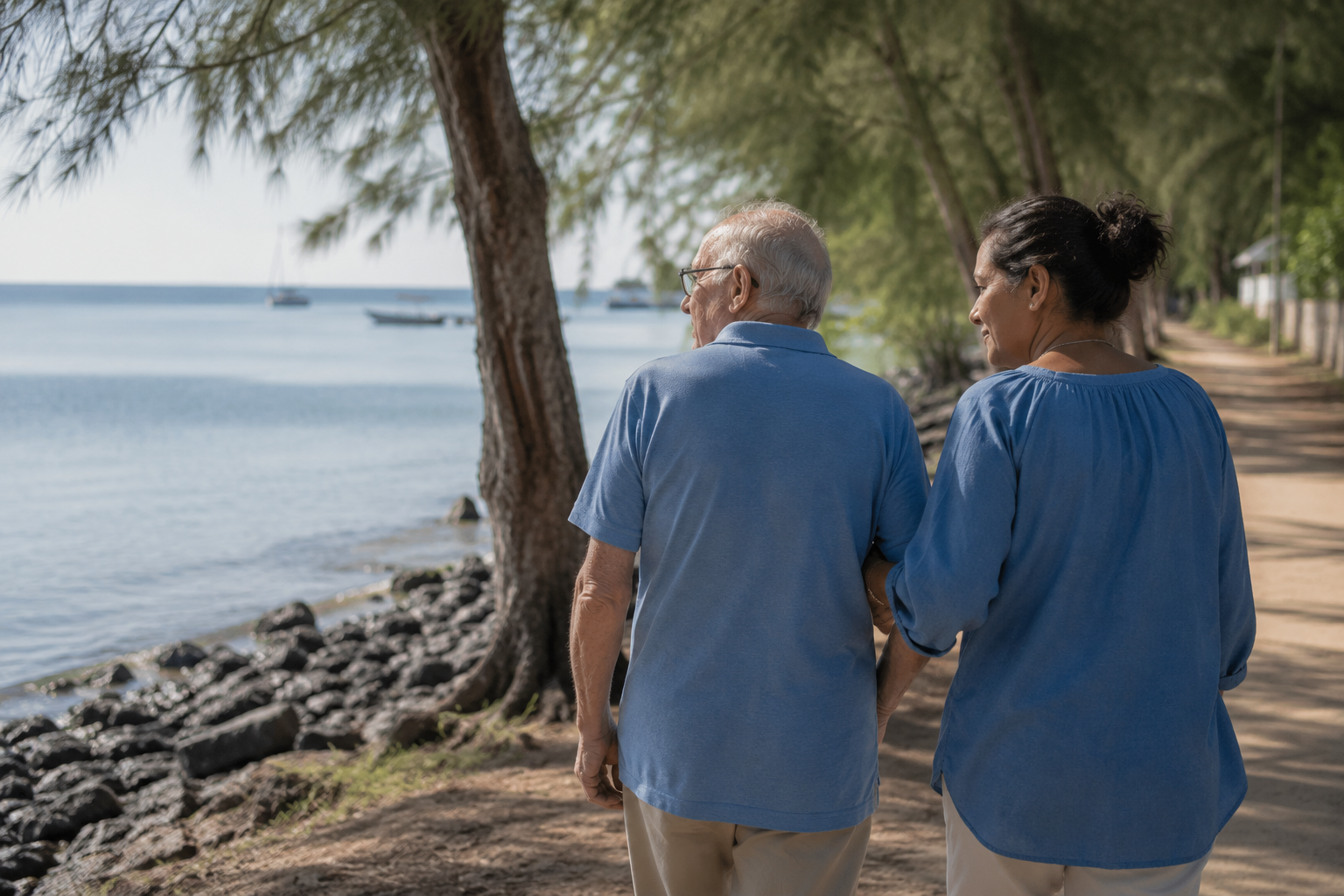 A carer walking arm in arm with an elderly man along a tree-lined coastal path in northern Mauritius