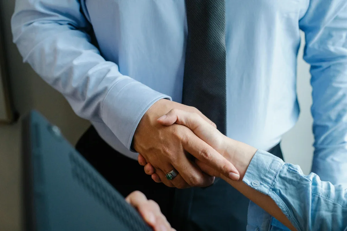 Two people shaking hands in a professional setting, one wearing a business suit and the other in a light blue shirt.