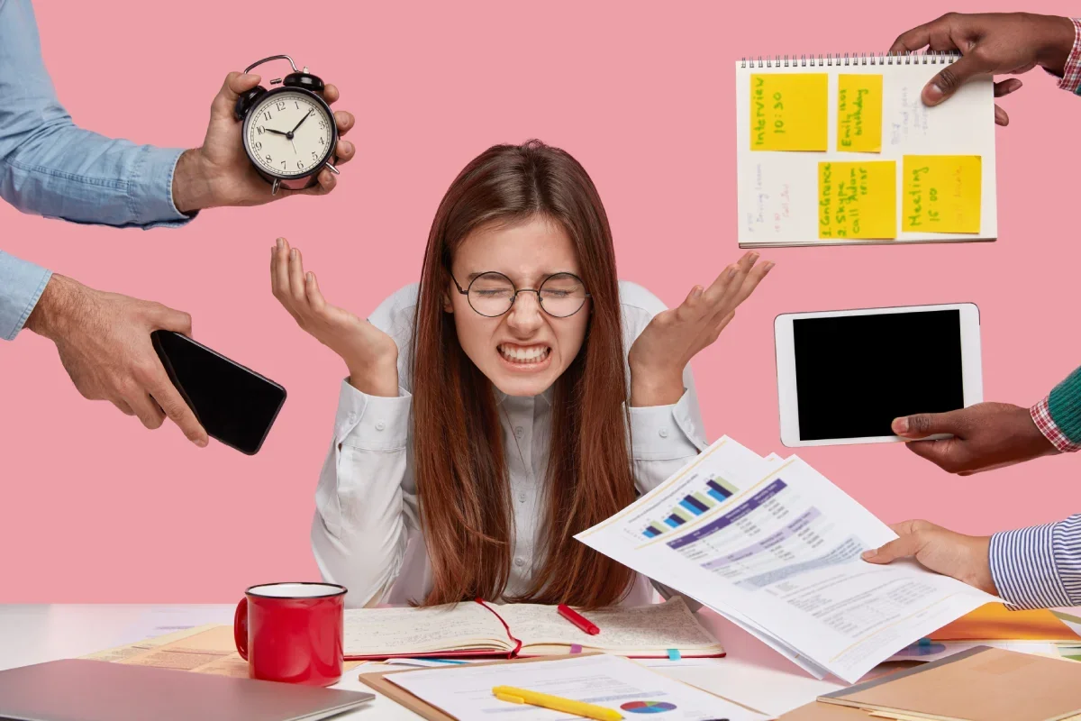 A woman with glasses looking stressed at a cluttered desk surrounded by multiple hands holding clocks, papers, a tablet, a smartphone, a planner, and sticky notes.