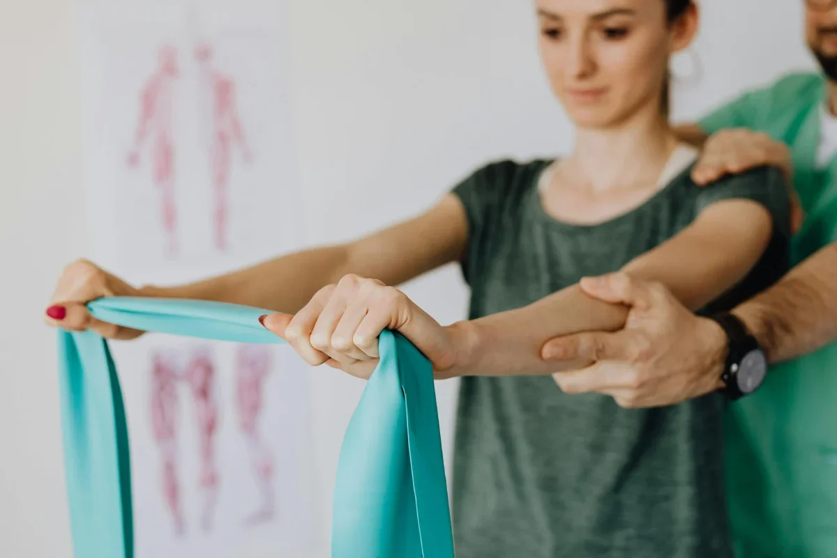 A physical therapy session where a woman is holding a resistance band, with an instructor assisting her, in a clinic with an anatomical chart in the background.