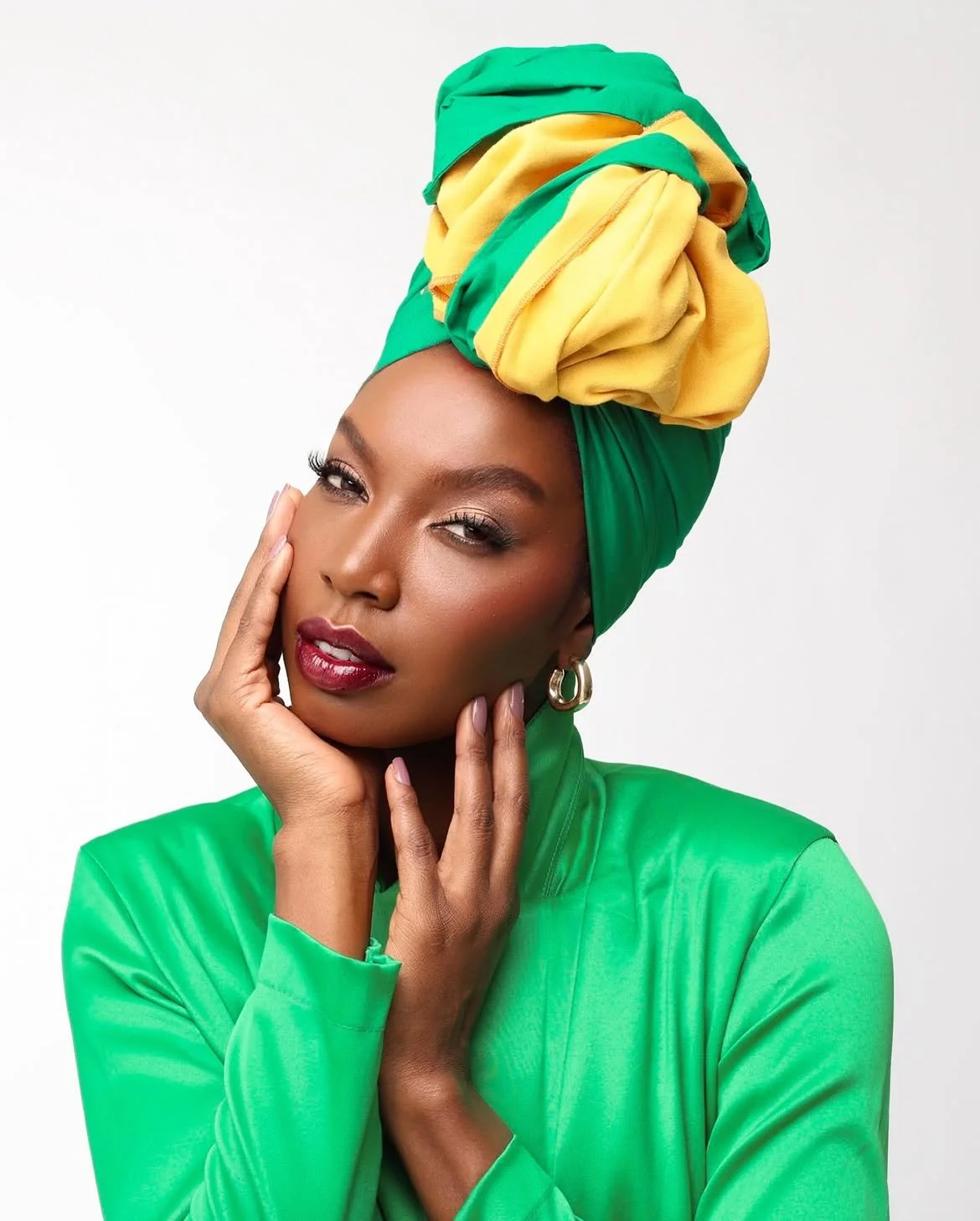 Mulher negra, Karina de Azevedo, usando turbante verde, detalhe em amarelo e roupa verde, segurando o rosto com uma expressão confiante.