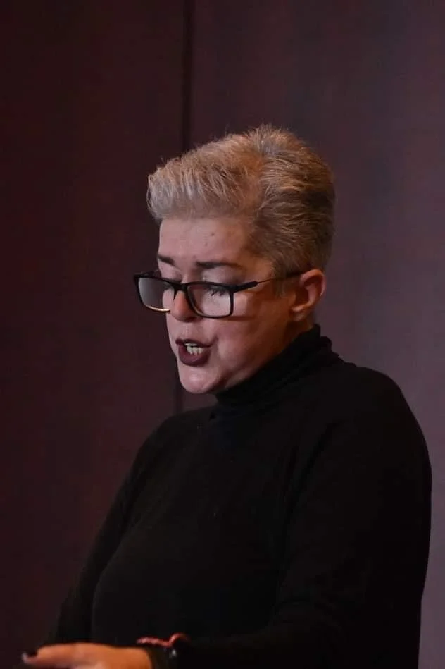 A woman with short light-colored hair, glasses, and dark lipstick speaking or reading against a dark background.
