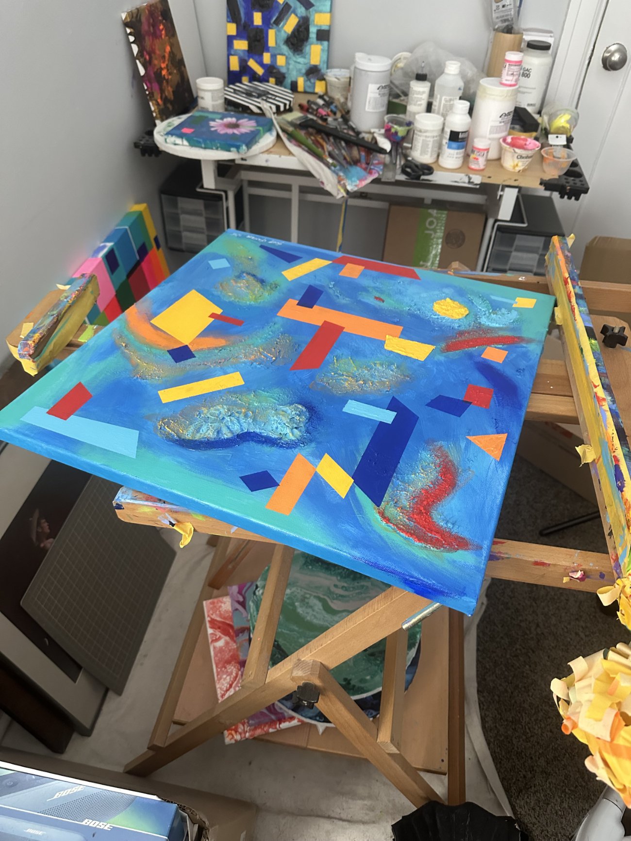 Colorful abstract painting on an easel in an artist's studio, surrounded by art supplies and completed artworks.