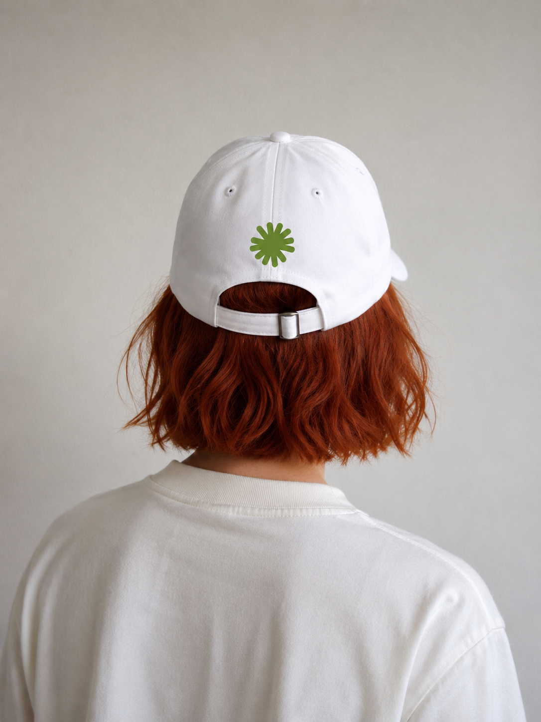 Back view of a person with shoulder-length red hair wearing a white baseball cap and a white sweatshirt. The cap has a green circular logo on the back.