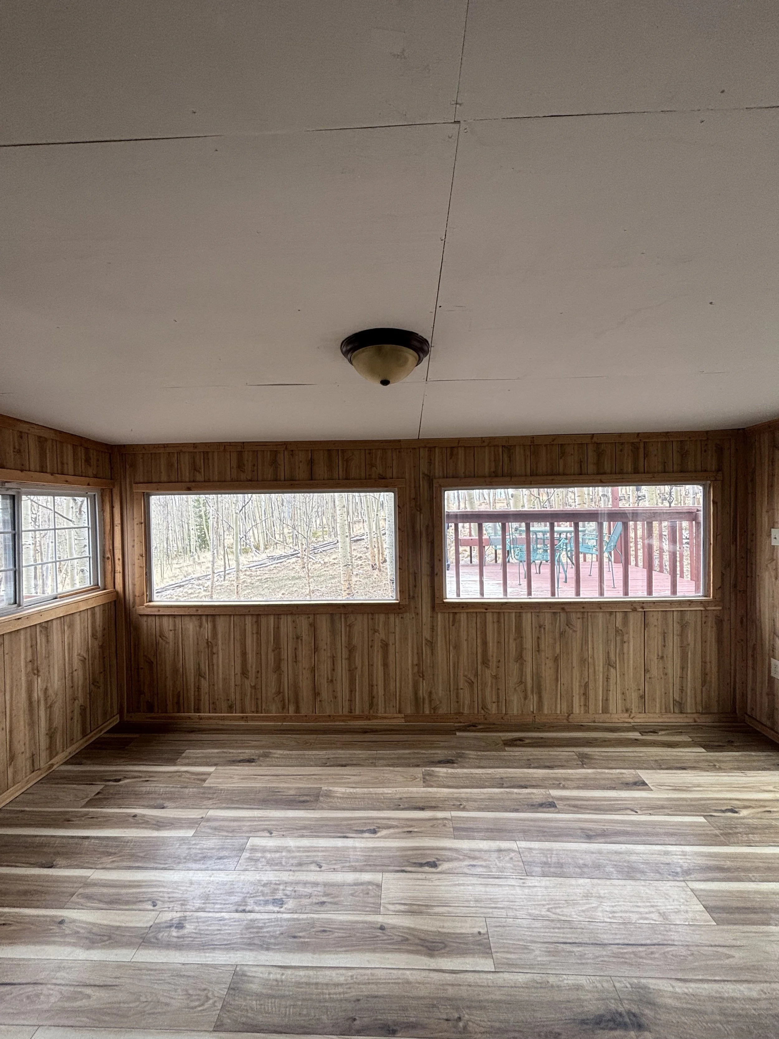 Empty sunroom with wood-paneled walls, large windows, and wood-look flooring, overlooking a deck with outdoor furniture.