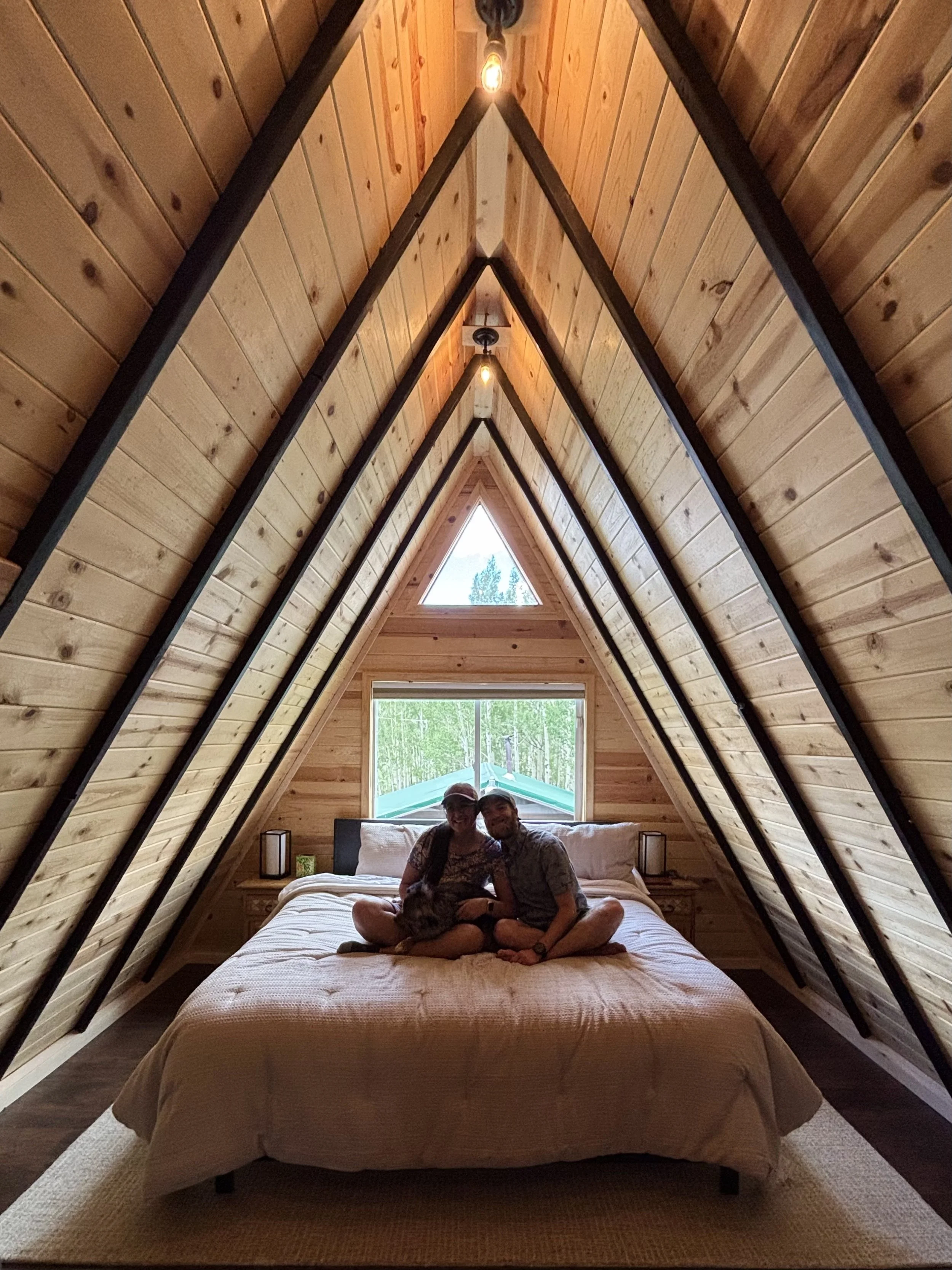 Two people sitting on a bed inside a wooden A-frame cabin loft, with triangular windows at the head of the bed and string lights hanging from the ceiling.