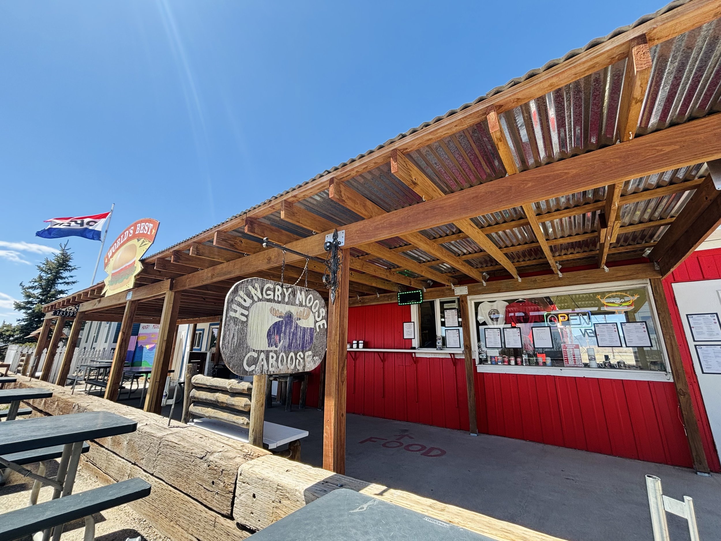 Outdoor food stand with a wooden roof, a sign for 'Hungry Moose Caboose,' and a flag flying above with a bright blue sky in the background.