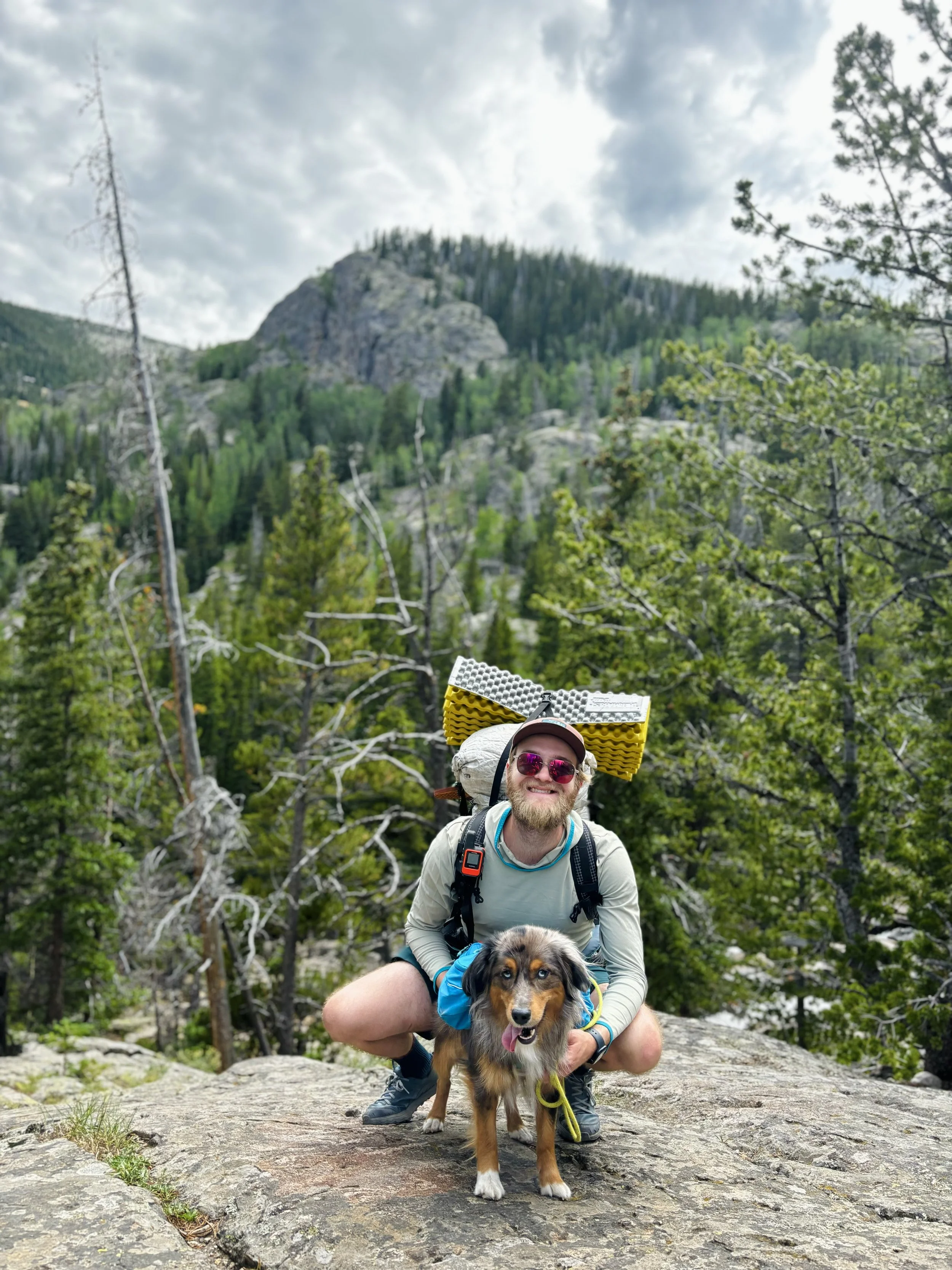 Nate and Acadia pose on a boulder in a forested mountain area with trees, rocks, and a cloudy sky in the background.