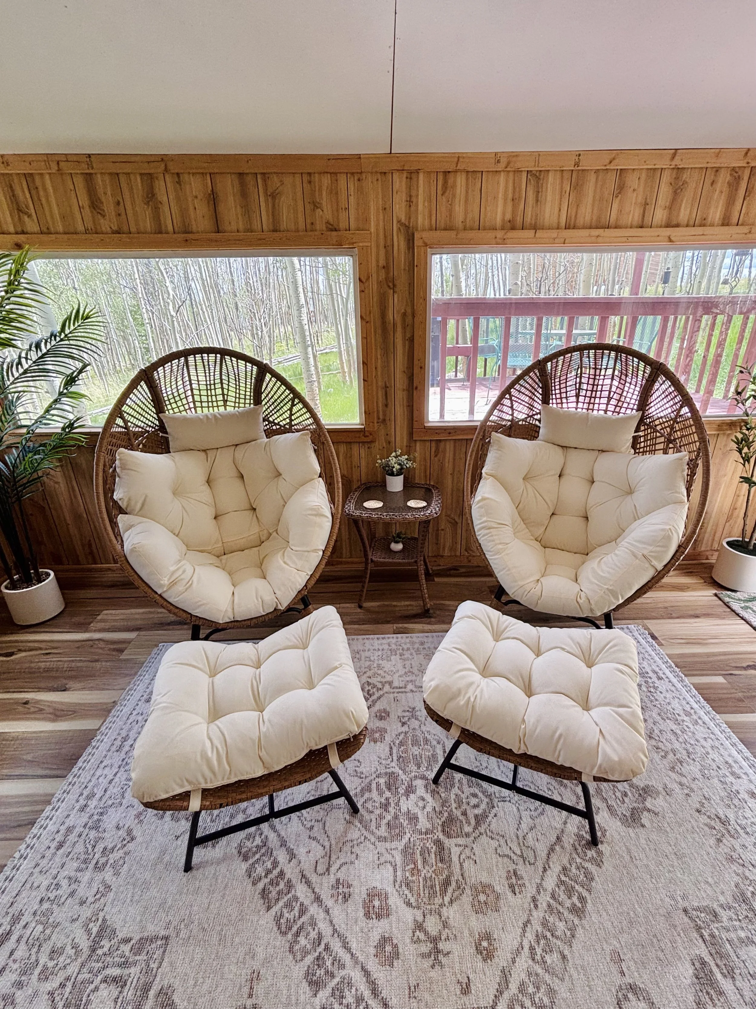 The image shows the sunroom with four cushioned chairs arranged around a small wooden side table with a small plant on it, all on a rug. The room has wooden walls and large windows revealing an outdoor view of trees and a wooden deck with a chair.
