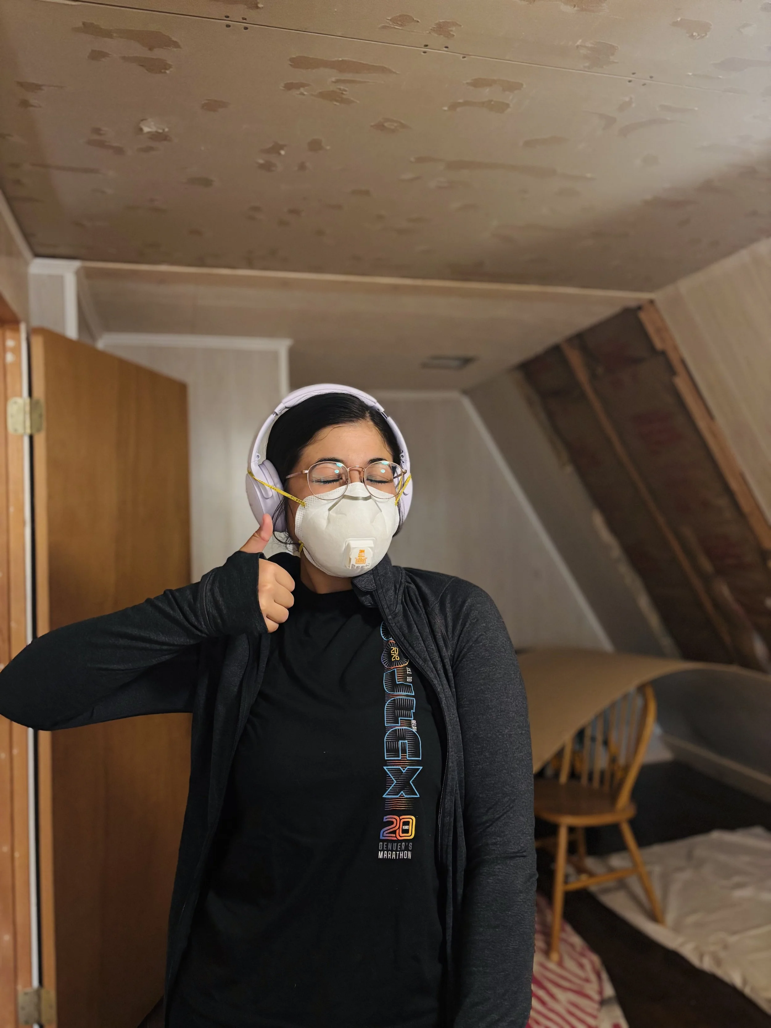 Leilani, wearing glasses and a protective face mask, gives a thumbs-up in an indoor setting with wooden chairs and a sloped ceiling in the background.