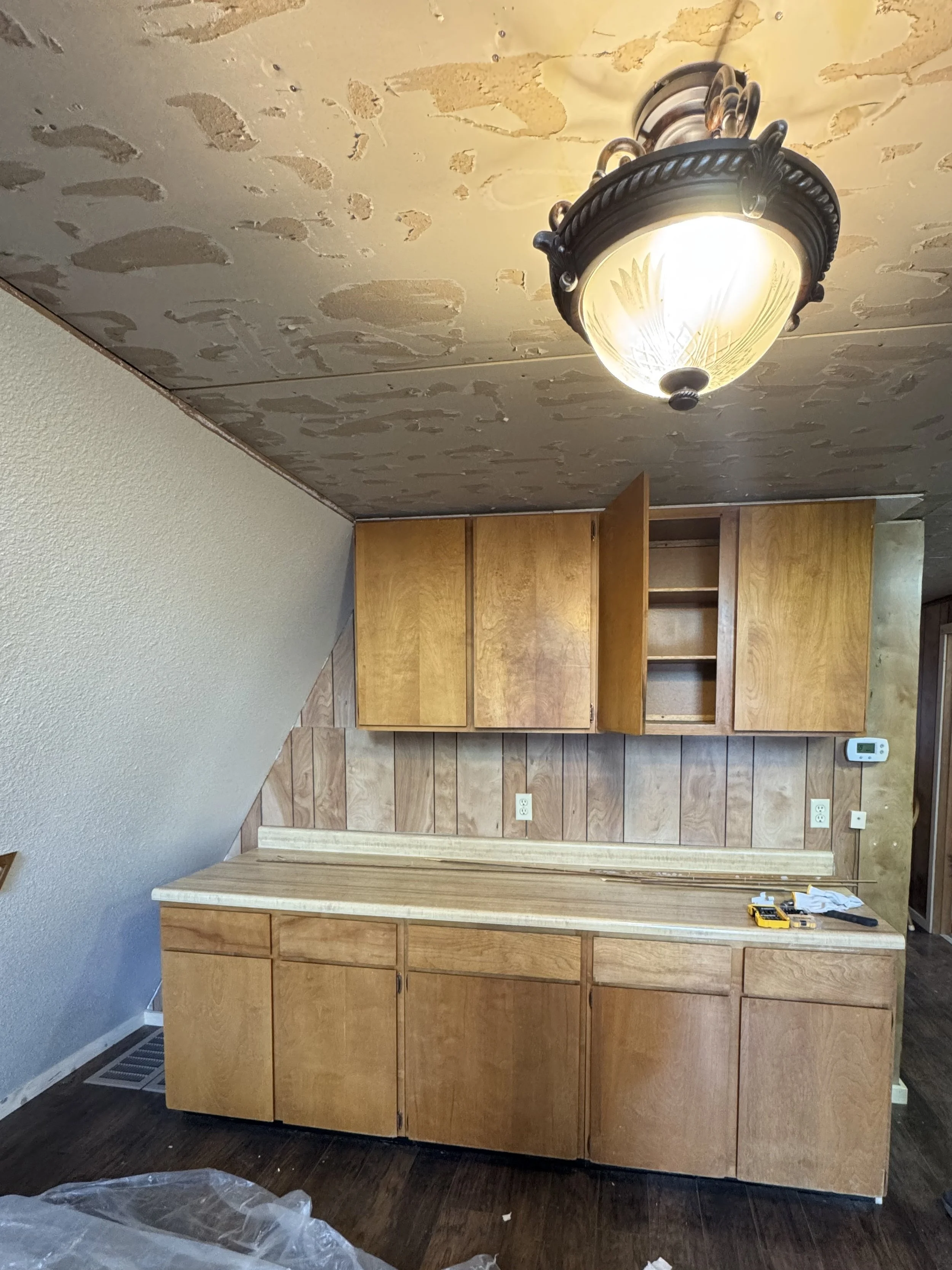 Kitchen cabinets with some doors open, tools on the countertop, ceiling with removed drywall patches, and a mounted light fixture.
