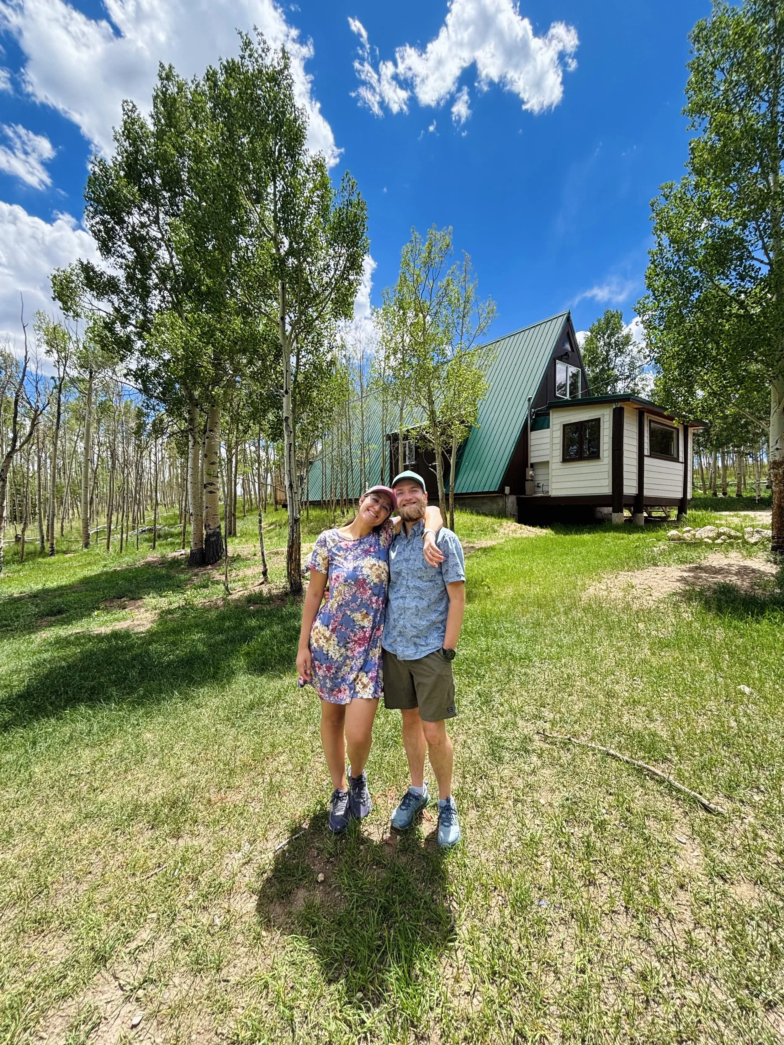 Leilani and Nate posing in front of the Kenosha Cabin, surrounded by trees and grass, under a partly cloudy blue sky.