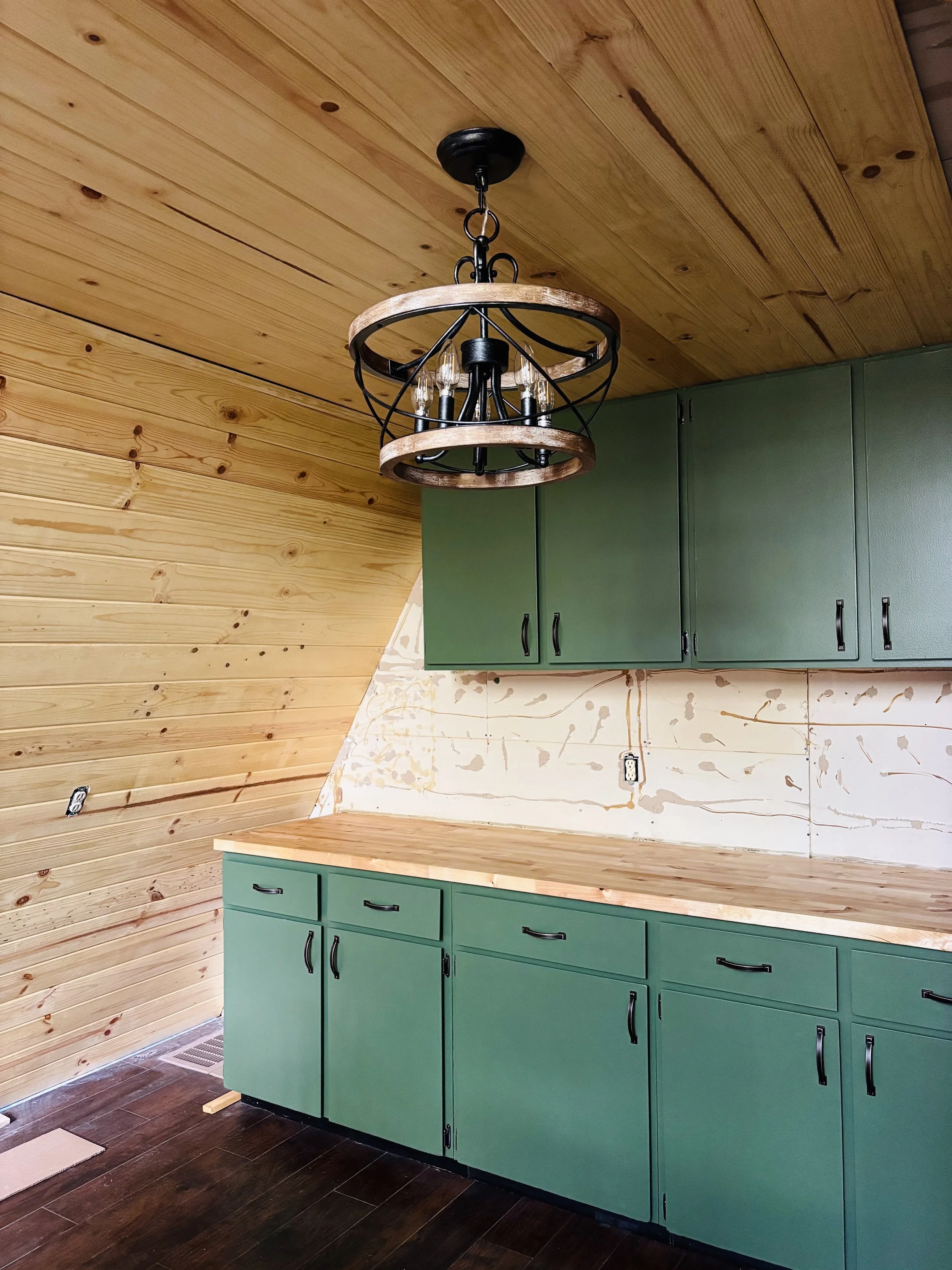 Interior of a kitchen under construction with green cabinets, wooden ceiling, and a hanging chandelier. The wall has marks and wiring for outlets.