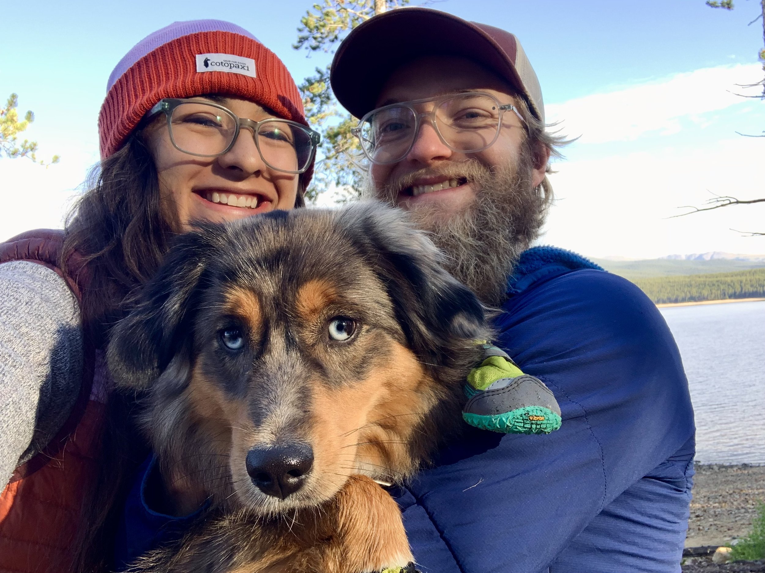 Leilani and Nate take a selfie outdoors while holding Acadia in front of them, with trees and water in the background on a sunny day.