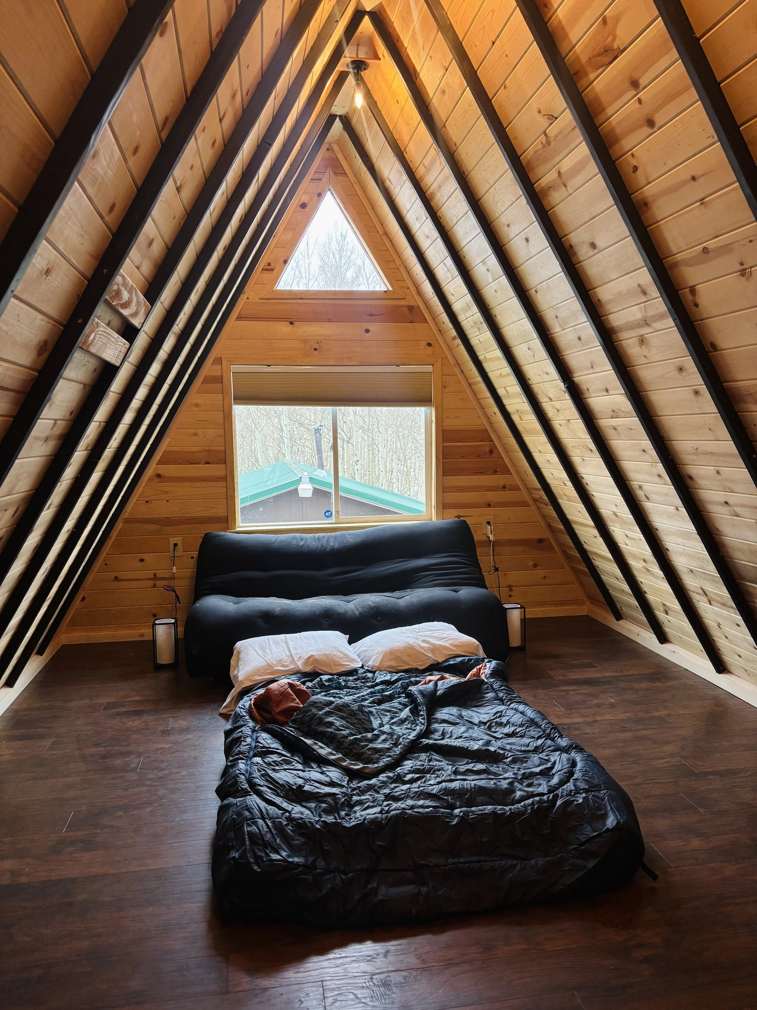 A cozy cabin loft with wood-paneled walls and ceiling. A black futon with white pillows is in the center of the room, and a sleeping bag is laid out on the floor in front of it.