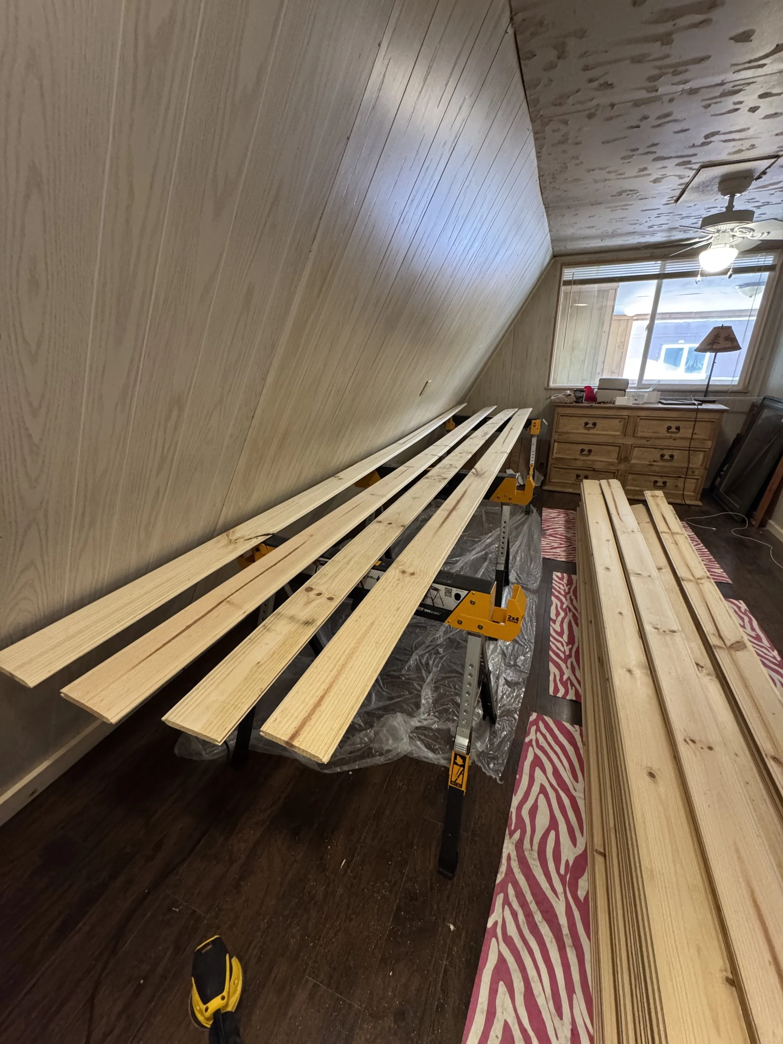 Interior of a room under renovation with wooden planks on black sawhorses, a dresser, a window with blinds, and a ceiling fan with light.