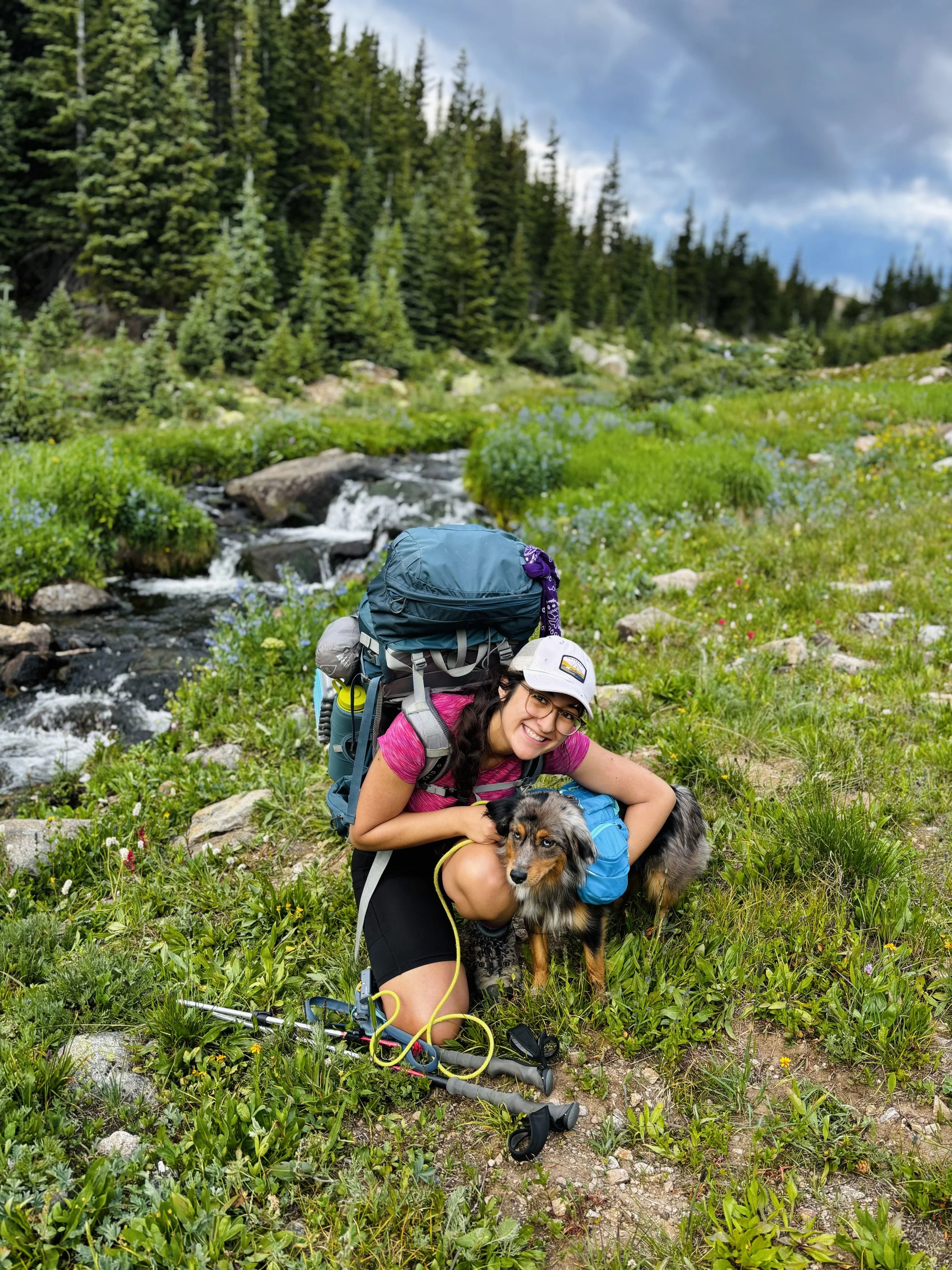 Leilani smiles while kneeling next to Acadia in a lush, green, mountainous area with a small stream, tall pine trees, and a cloudy sky.