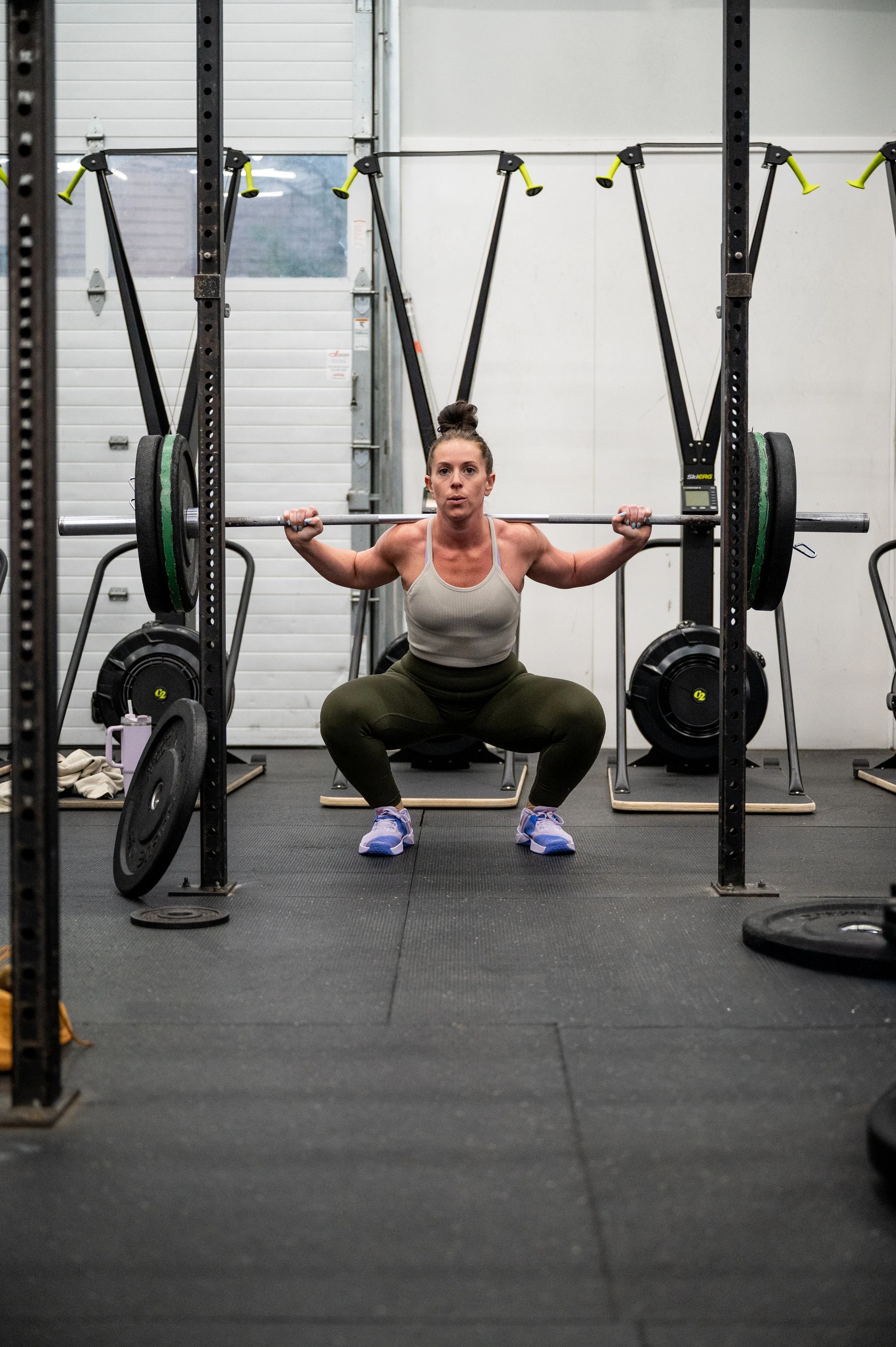 A woman is performing a squat exercise with a barbell loaded with weight plates on her shoulders inside a gym. She is wearing a gray top, black tights, and purple sneakers. 