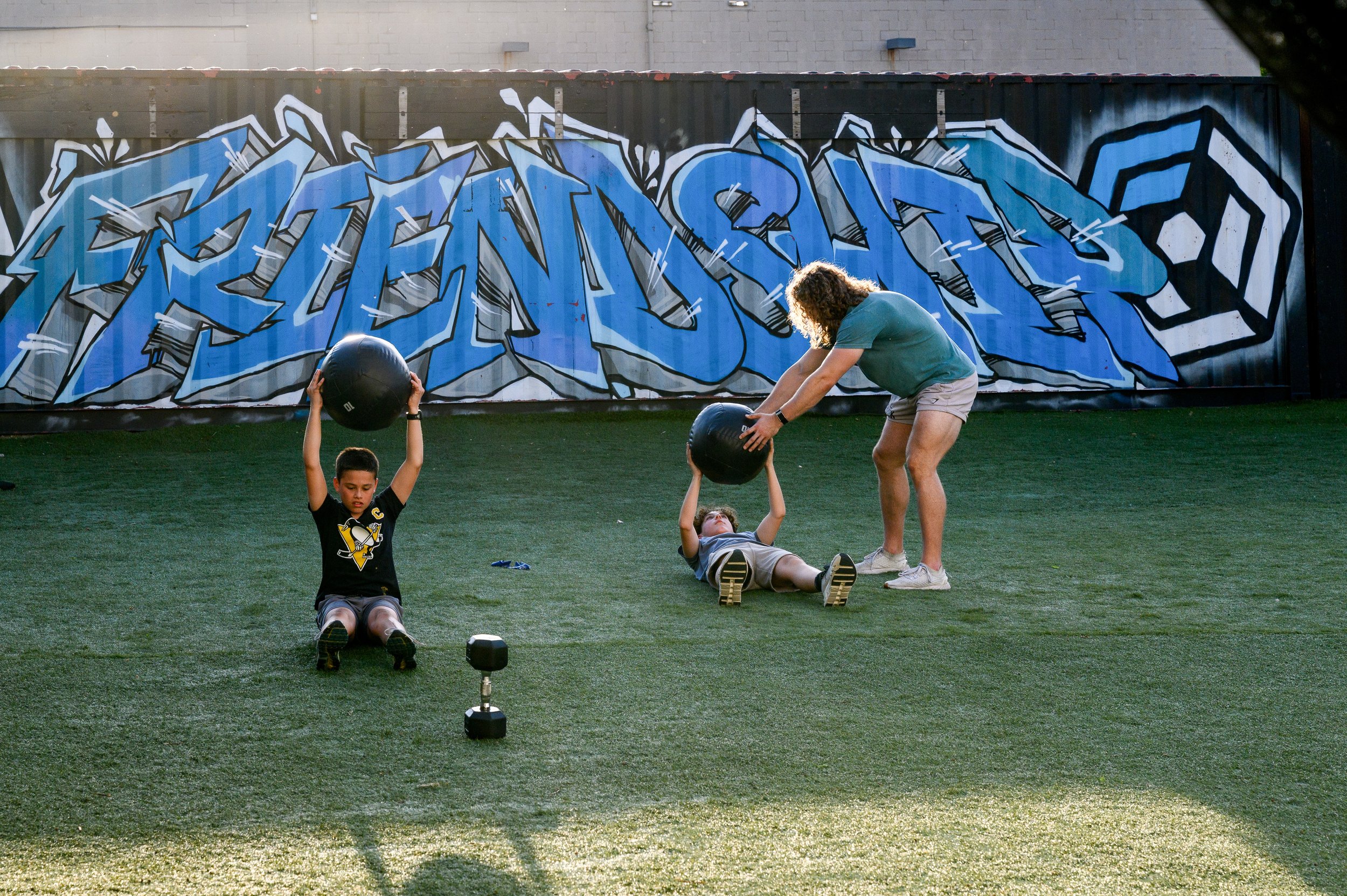 People exercising outdoors on a grassy field with a graffiti-covered wall in the background. A woman appears to be instructing children in a fitness activity with weighted balls.