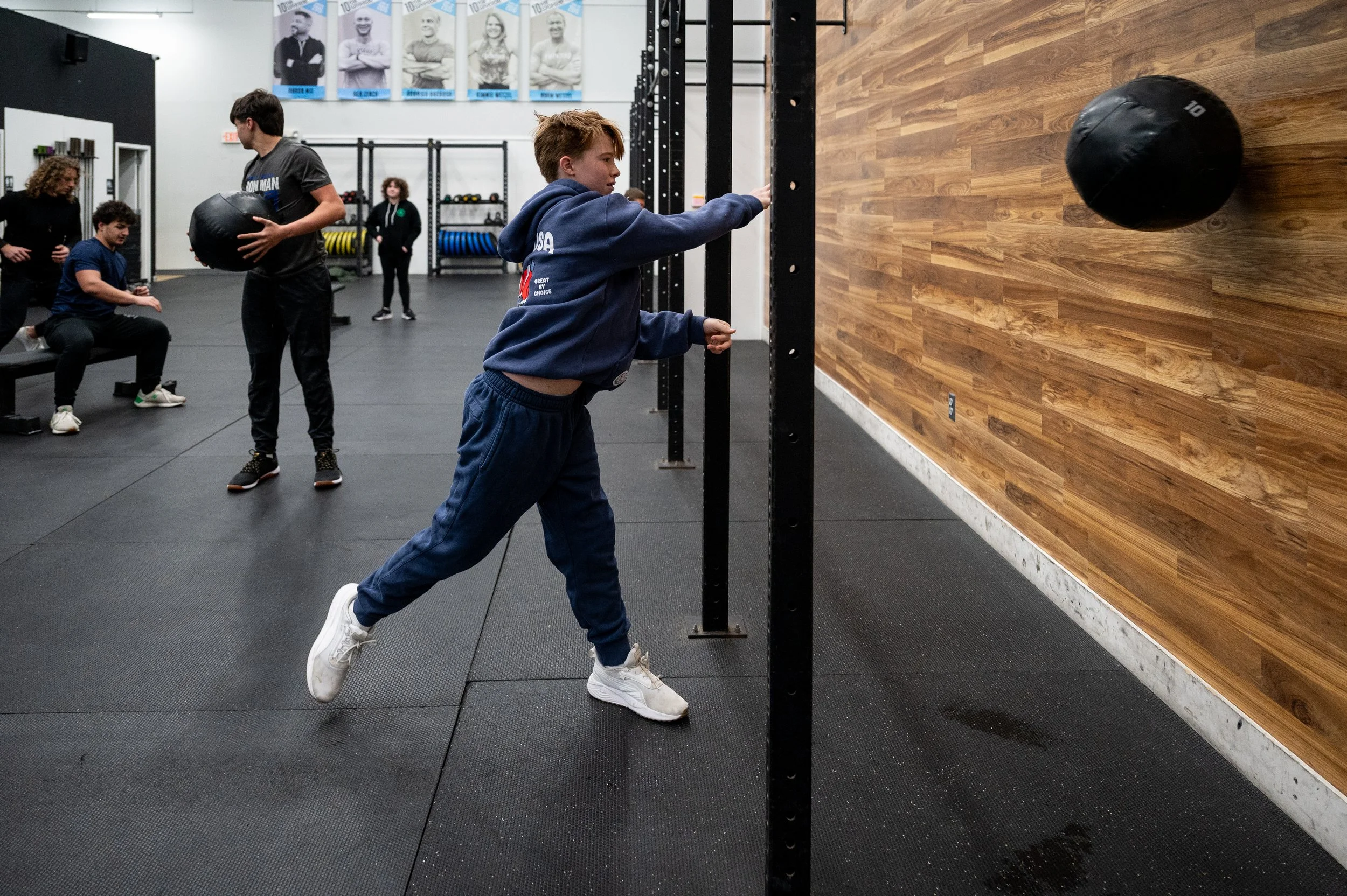 A young boy wearing a blue hoodie and blue pants throws a black medicine ball against a wooden wall in a gym. There are several other people in the gym, including a seated woman, a man holding a medicine ball, and other individuals in the background 