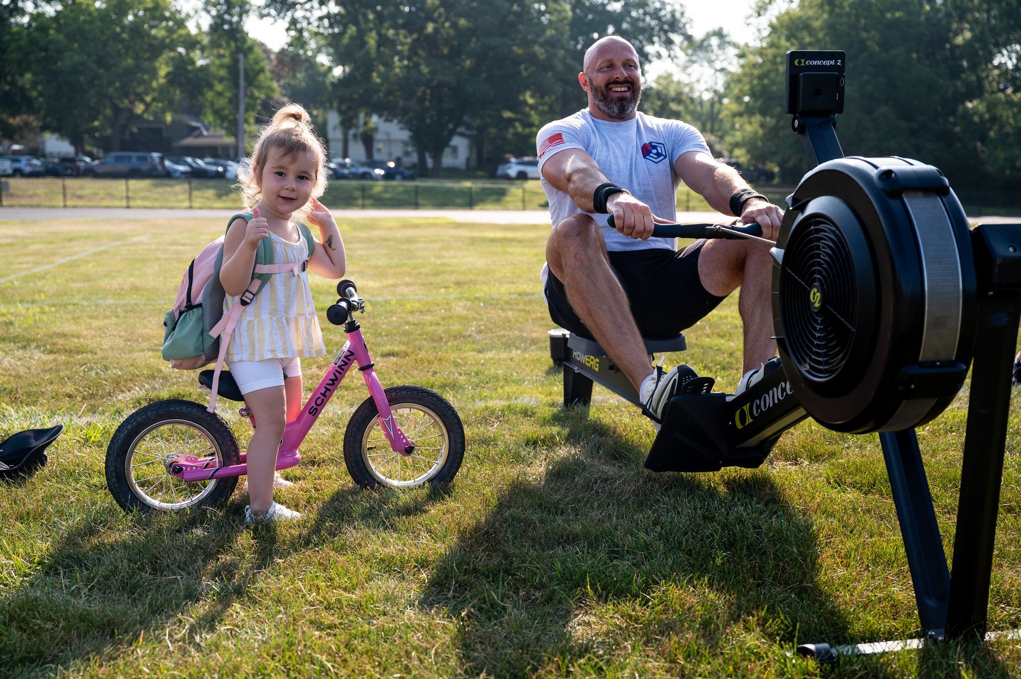 A man and a young girl at a park. The girl is on a pink bicycle with training wheels, giving a thumbs up. The man is pedaling a stationary exercise bike, smiling. Both are outdoors on grass, with trees and parking lot in the background.