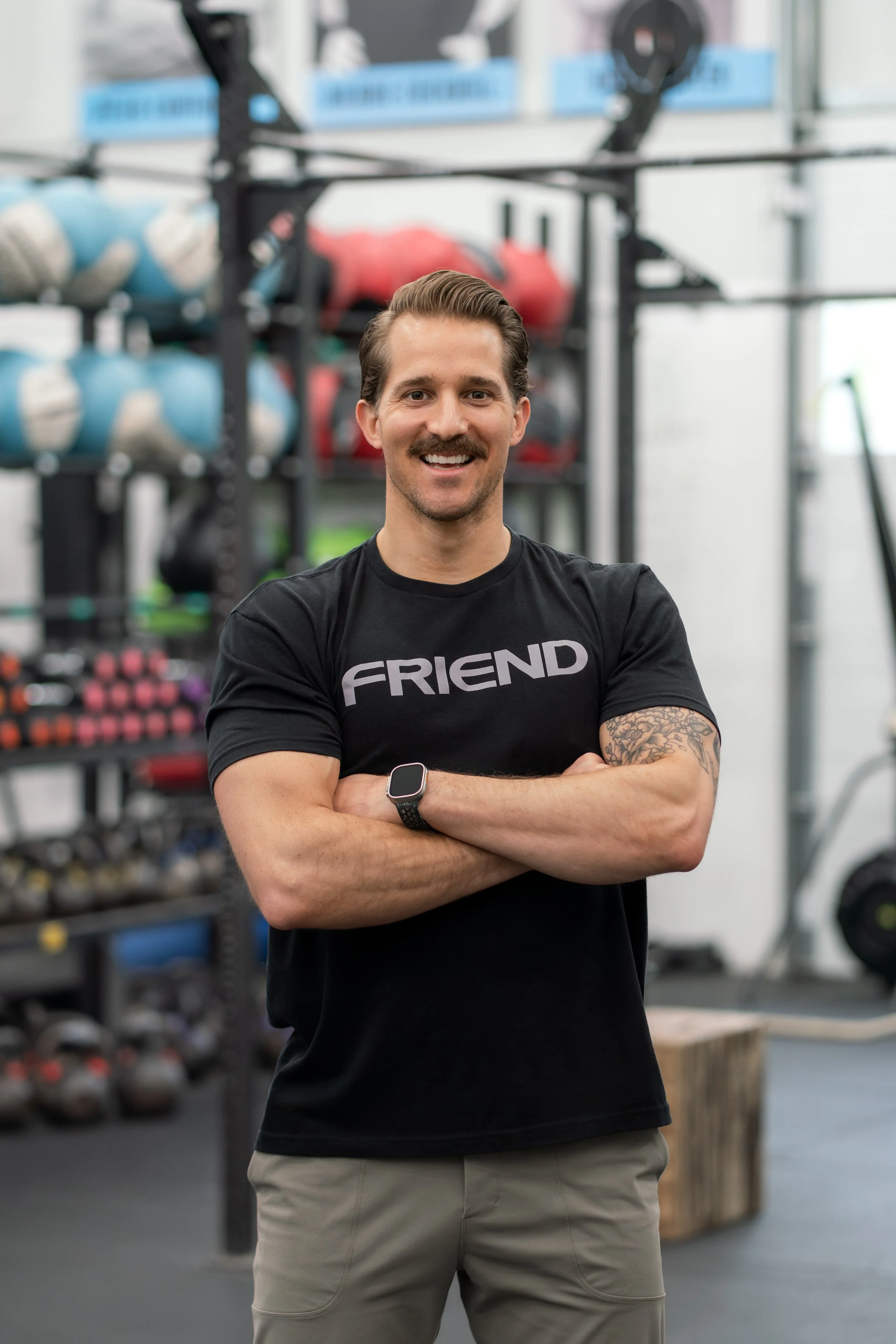 A smiling man with a mustache and tattoo on his left arm standing with arms crossed in a gym, wearing a black t-shirt with the word 'FRIEND' on it, with fitness equipment in the background.