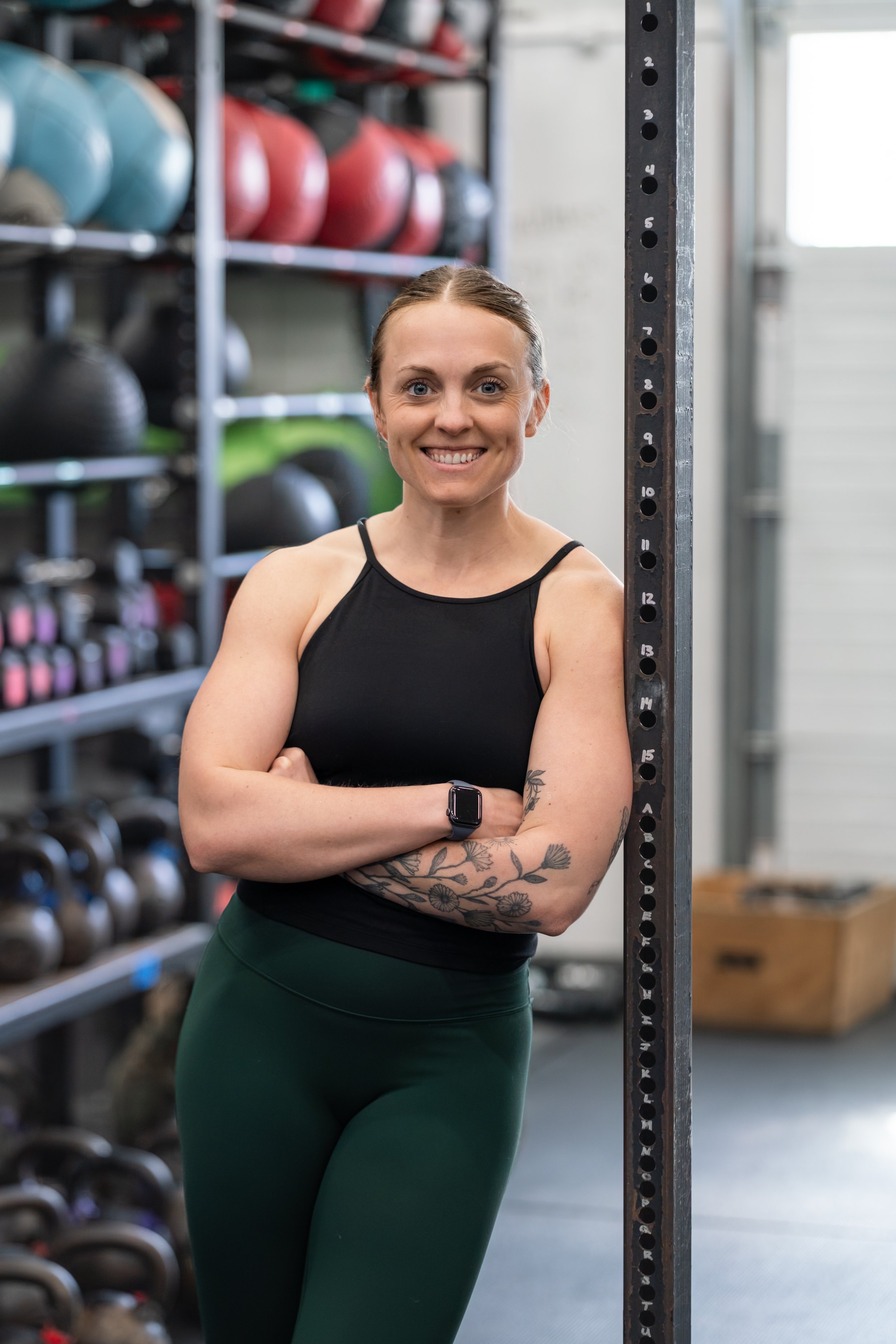 A smiling woman with tattoos on her arm stands in a gym, leaning against a black steel squat rack, with shelves filled with colorful medicine balls in the background.