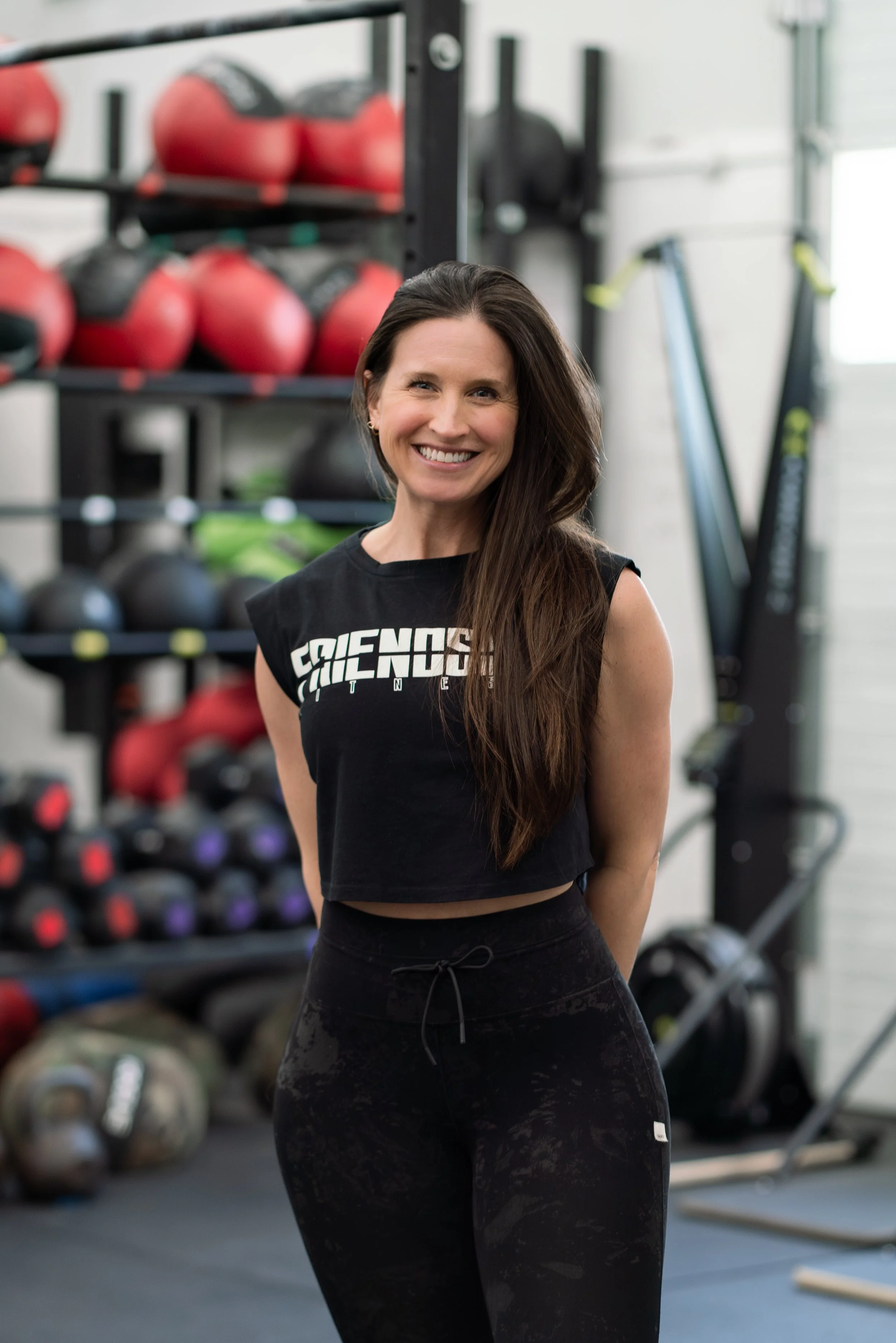 A smiling woman with long dark hair in a gym standing in front of a rack of kettlebells and weight plates.