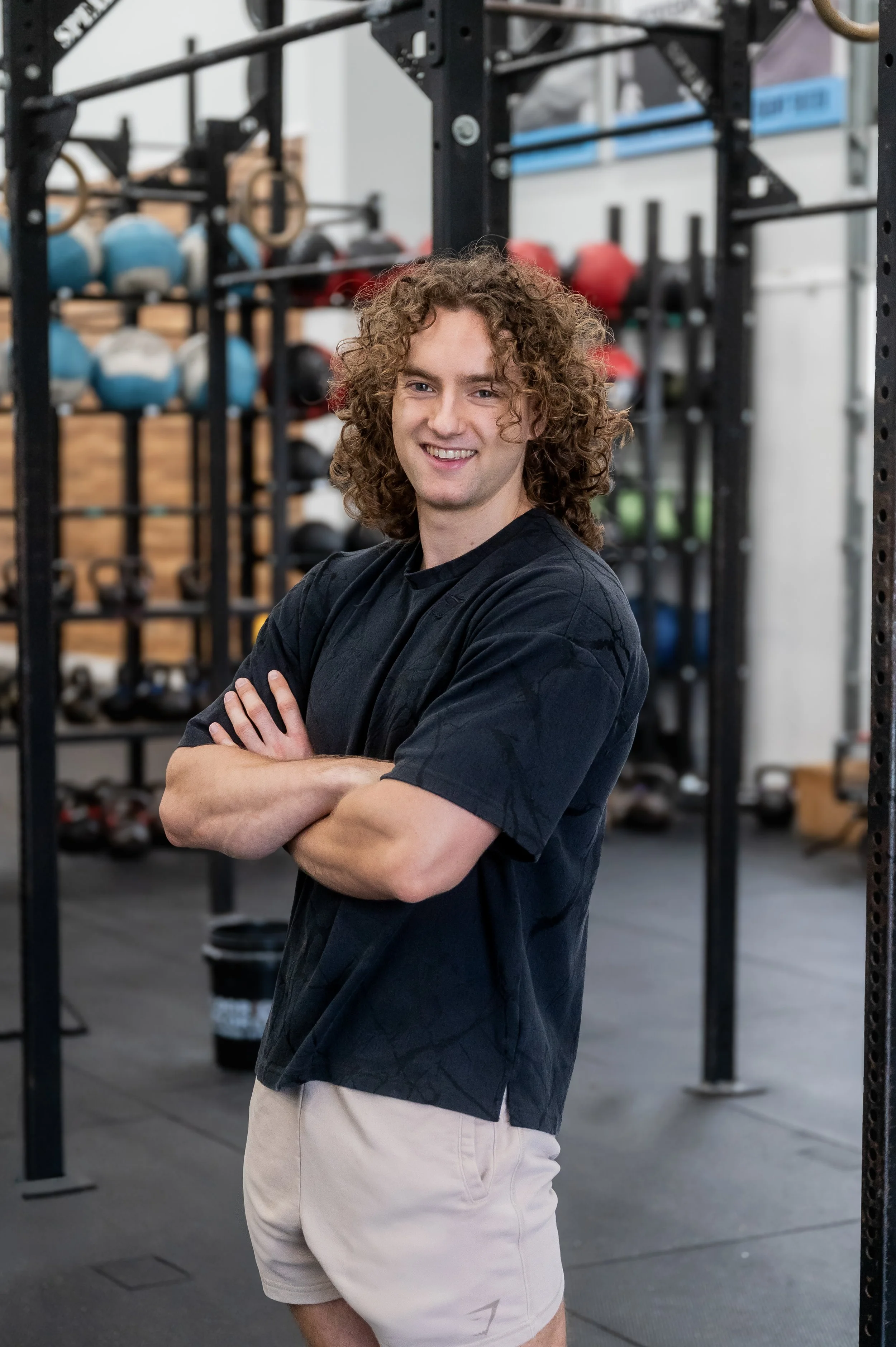 A smiling man with curly hair wearing a black shirt and beige shorts standing in a gym with black metal racks and workout equipment in the background.